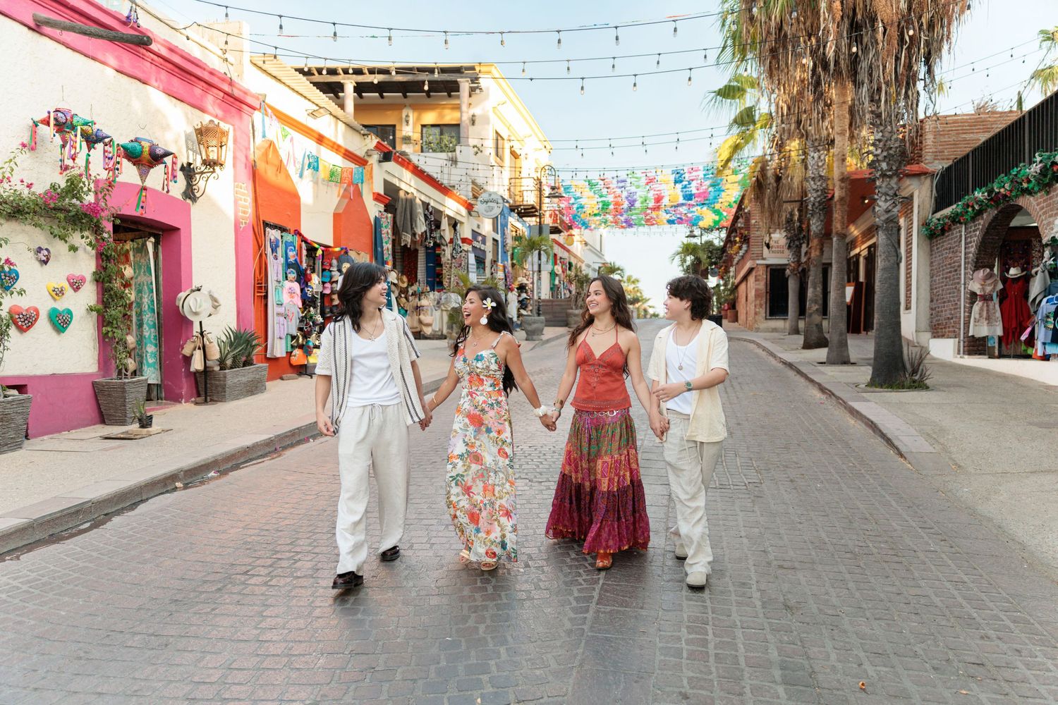 Four women walk hand-in-hand down a colorful Mexican street market decorated with festive papel picado banners.