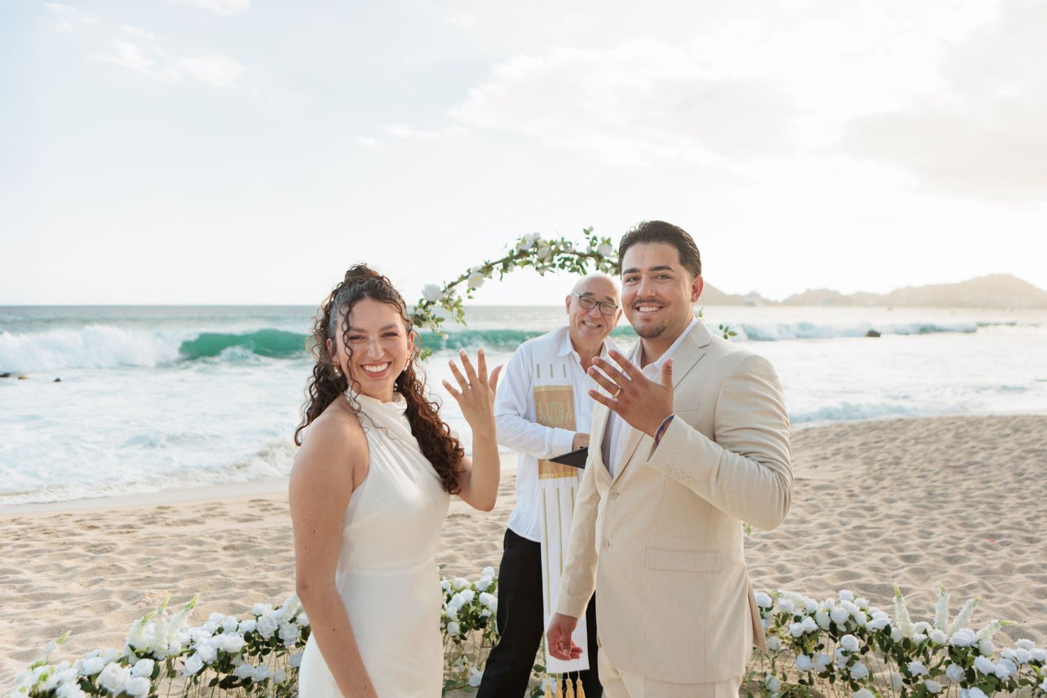 Couple showing wedding rings on a beach ceremony with floral arch and ocean waves in the background.