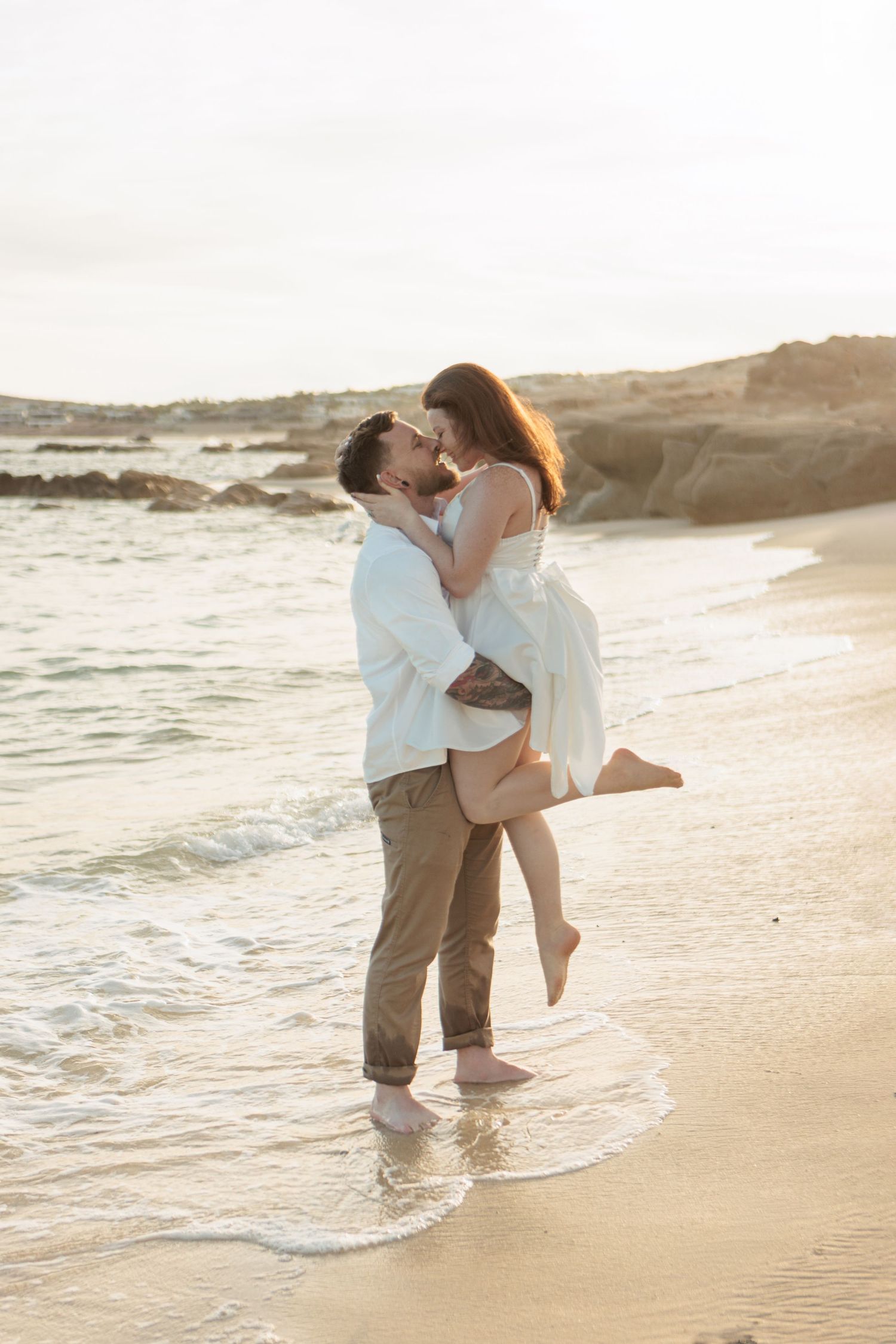Romantic couple kissing on beach shoreline at sunset, man lifting woman in white dress.