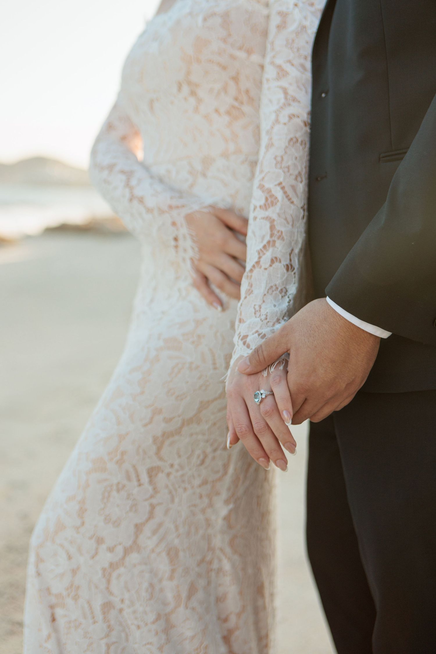 Close-up of bride and groom holding hands on beach, showcasing diamond engagement ring and lace dress.