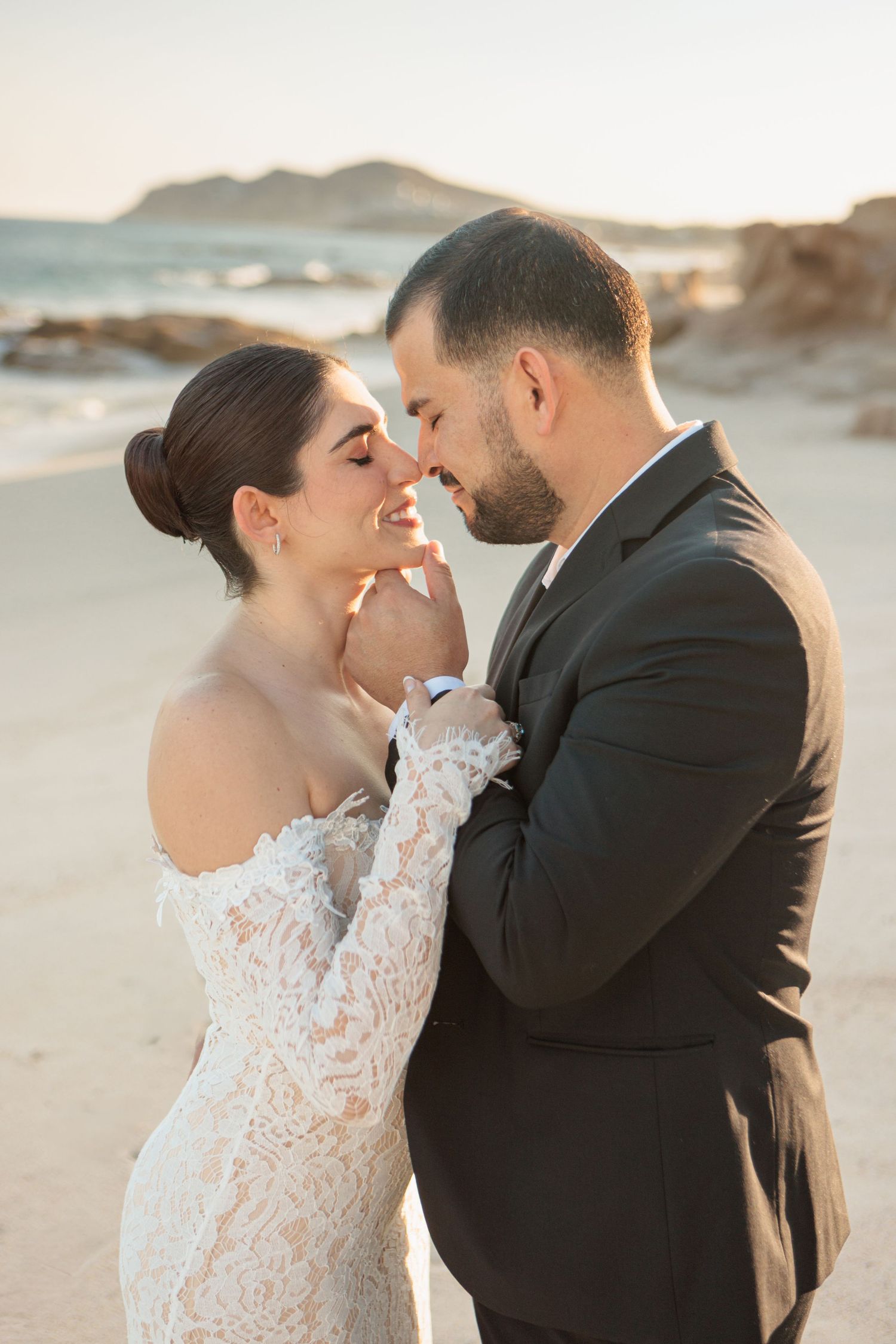 Bride in lace off-shoulder gown and groom in dark suit share romantic moment on sandy beach at golden hour.
