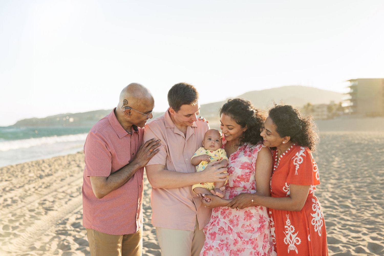 Multigenerational family smiling together on a sunny beach, admiring a newborn baby held by parents.