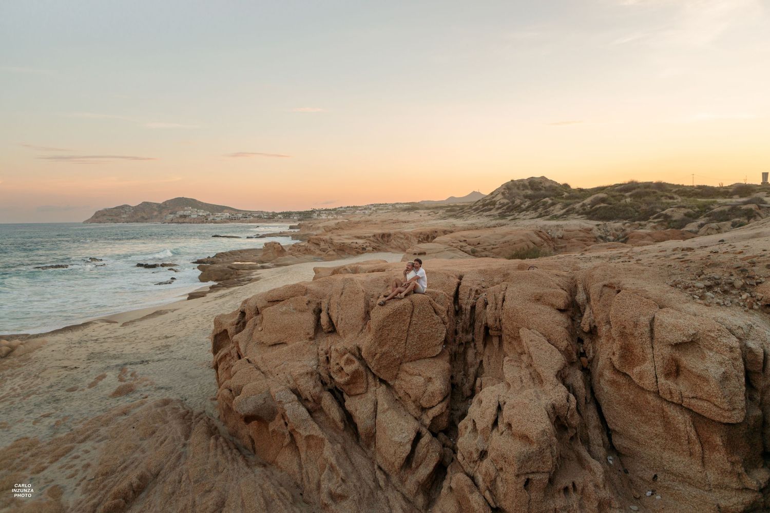 Person sitting on rocky coastal cliffs at sunset with ocean and hills in background.