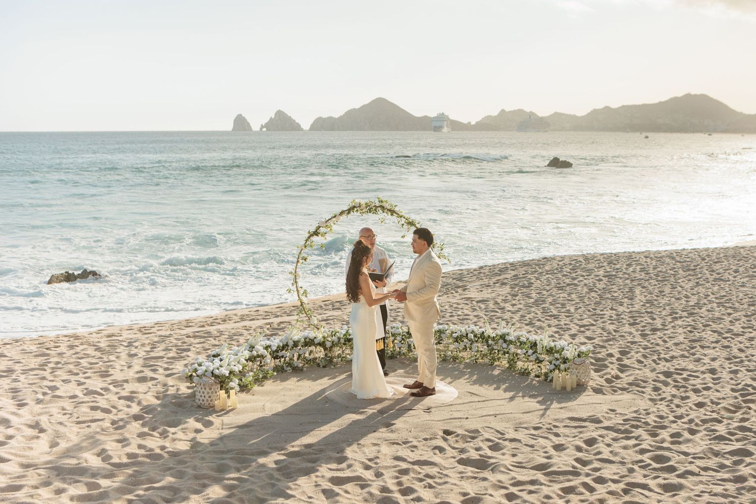 Couple exchanging vows at a beach wedding ceremony with a floral arch and ocean backdrop.