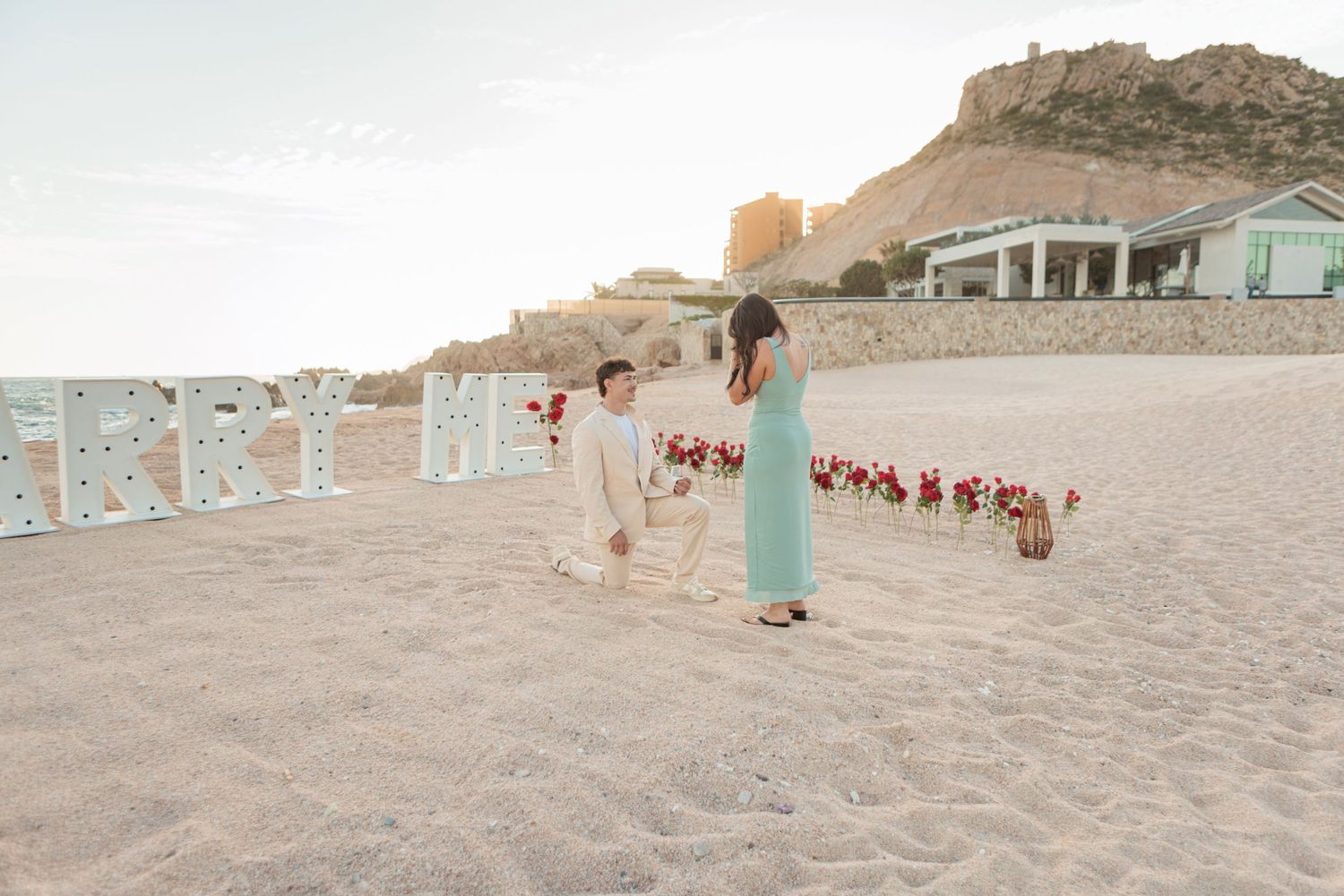 Man proposing to woman on beach with floral arrangement and marquee letters at sunset.