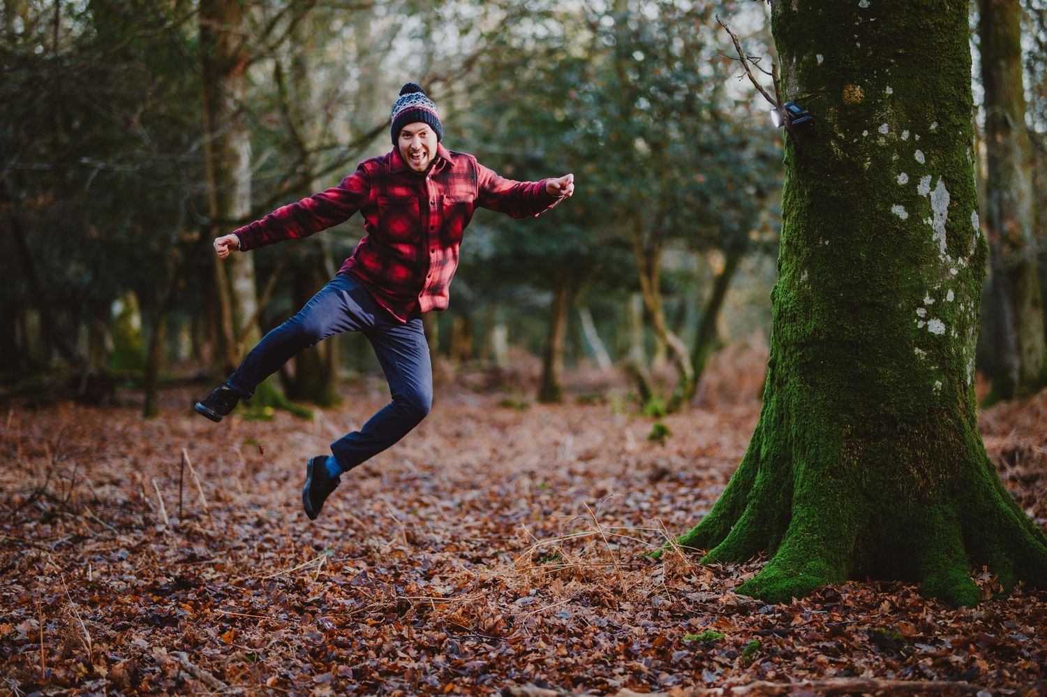 Person in red jacket and jeans jumps playfully through autumn forest covered in fallen leaves.