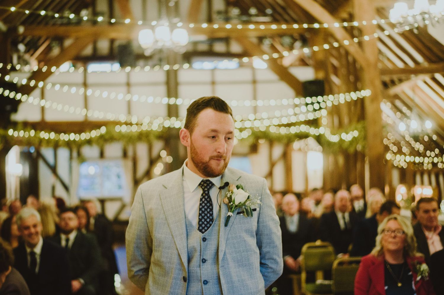 A wedding ceremony takes place in a rustic barn venue decorated with twinkling string lights and floral arrangements.