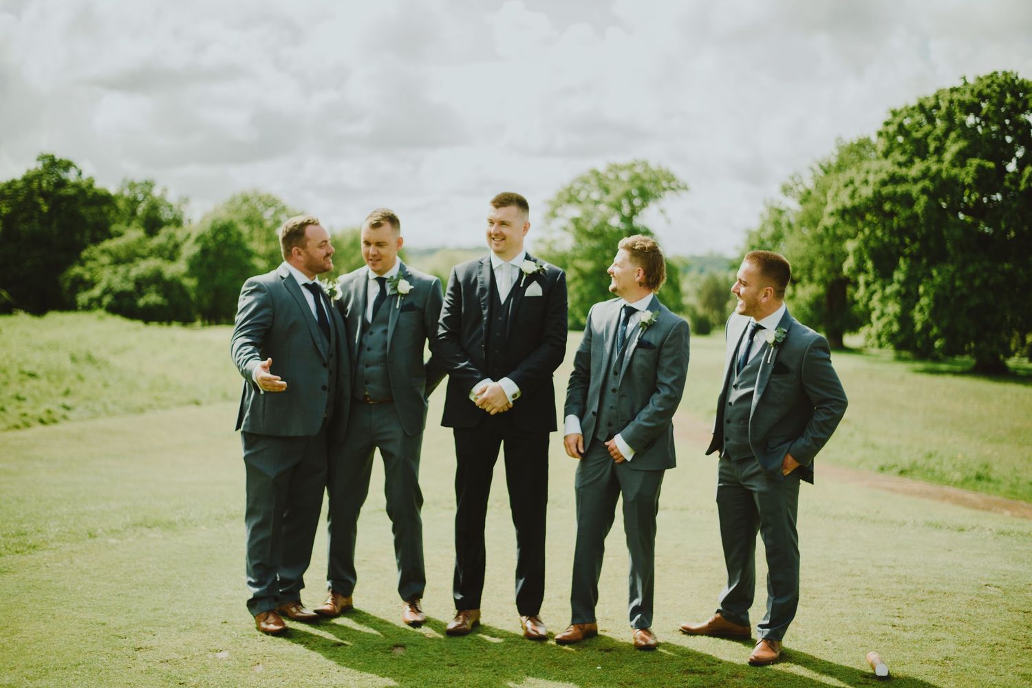 Wedding party in dark suits walking in a line on a grassy hill on an overcast day.