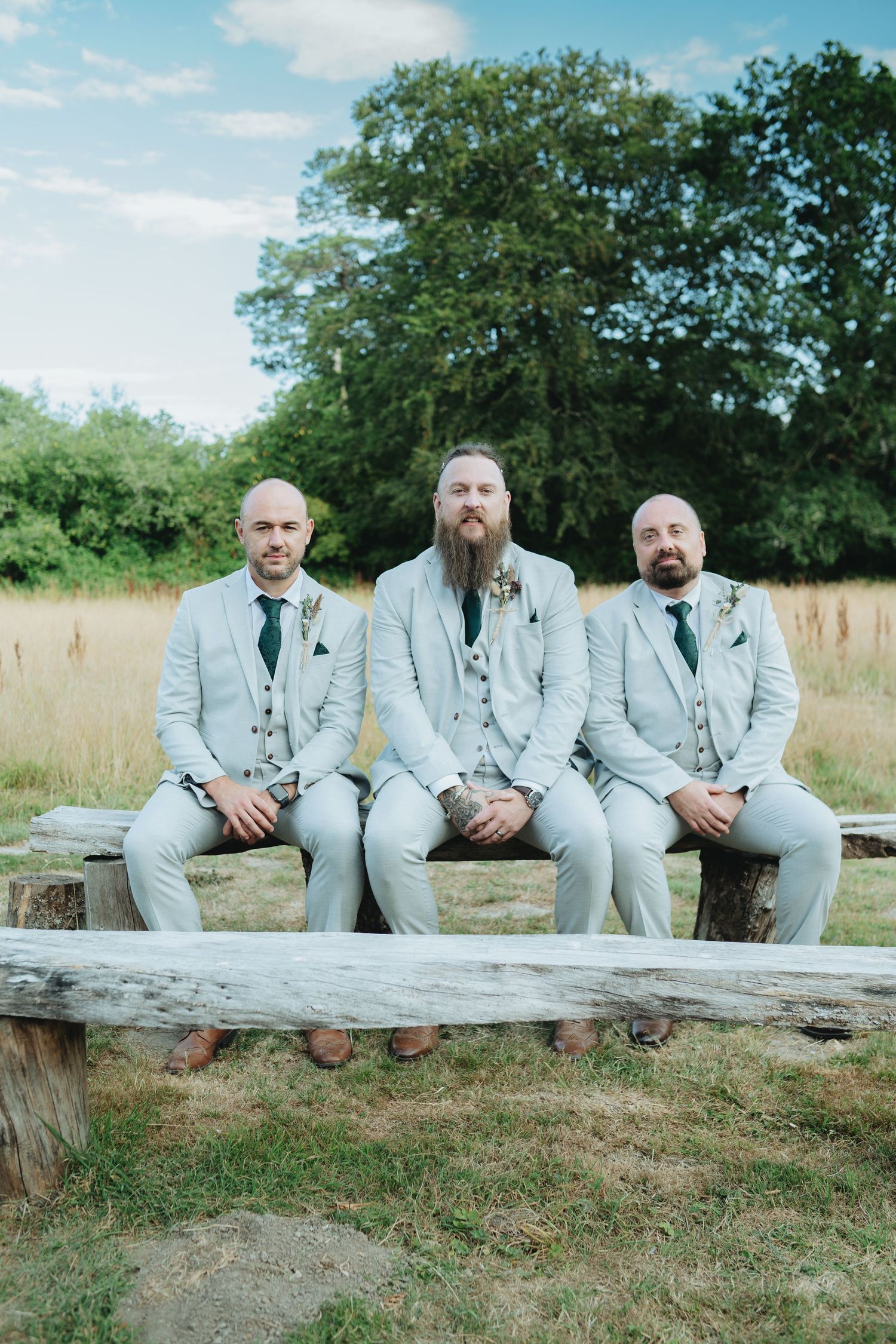 Three groomsmen in light grey suits sit on a rustic wooden bench outdoors at a wedding.