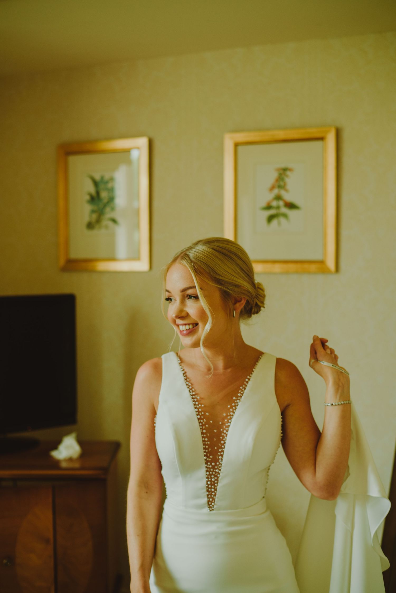 A bride in an elegant white wedding dress with beaded neckline poses in front of framed botanical artwork.