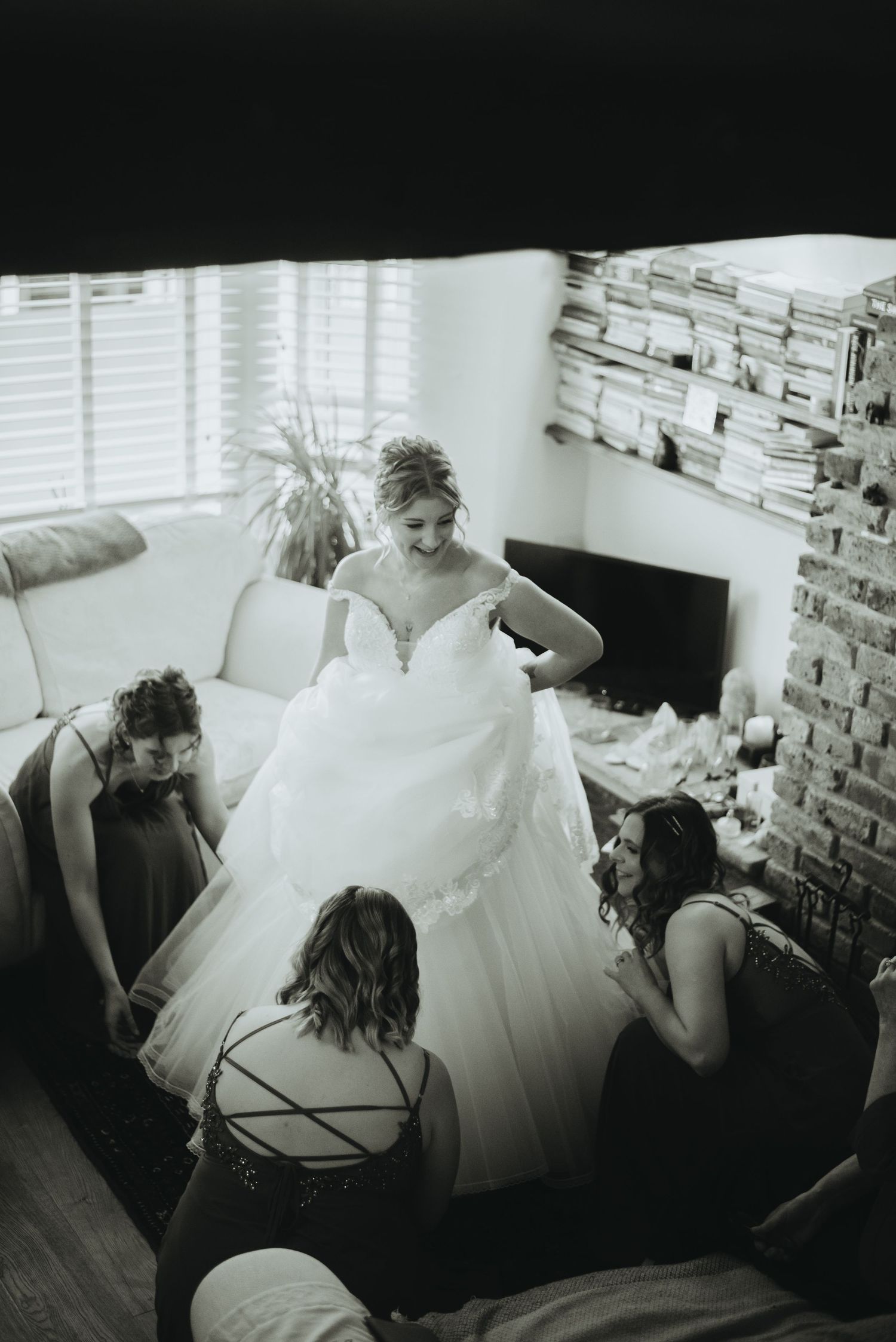 Black and white photo of a bride in a ball gown surrounded by bridesmaids helping her prepare in a cozy living room.