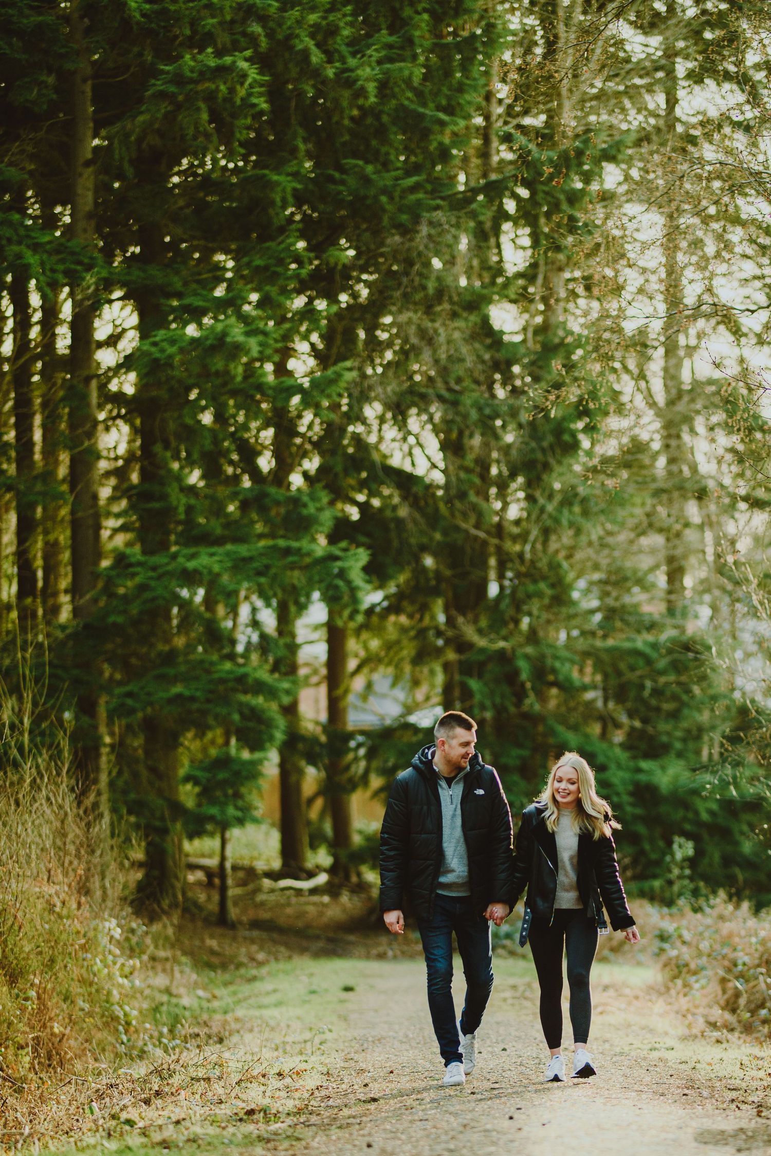 A couple walks along a forested path in autumn, with sunlight filtering through evergreen trees.