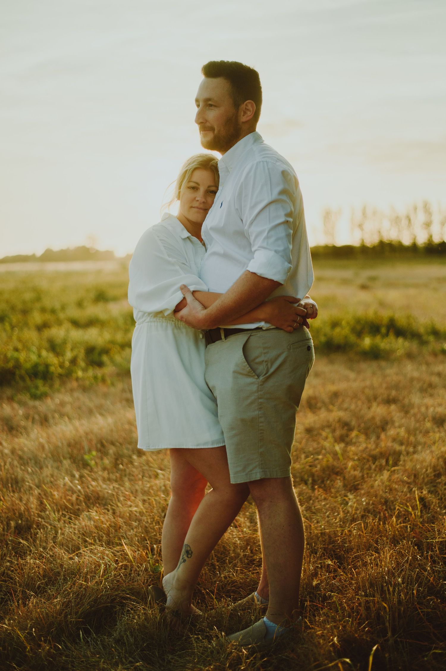 A couple embraces during golden hour in a sunlit field while wearing light-colored casual clothing.