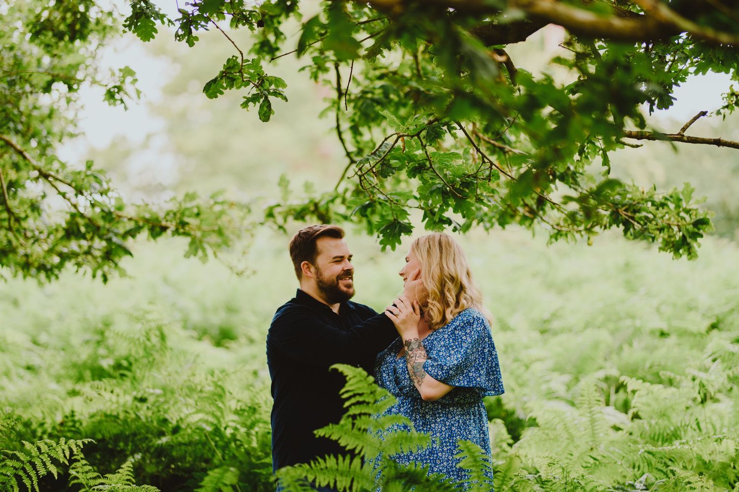A couple holds hands and leans together during a romantic photoshoot in a lush green garden setting.