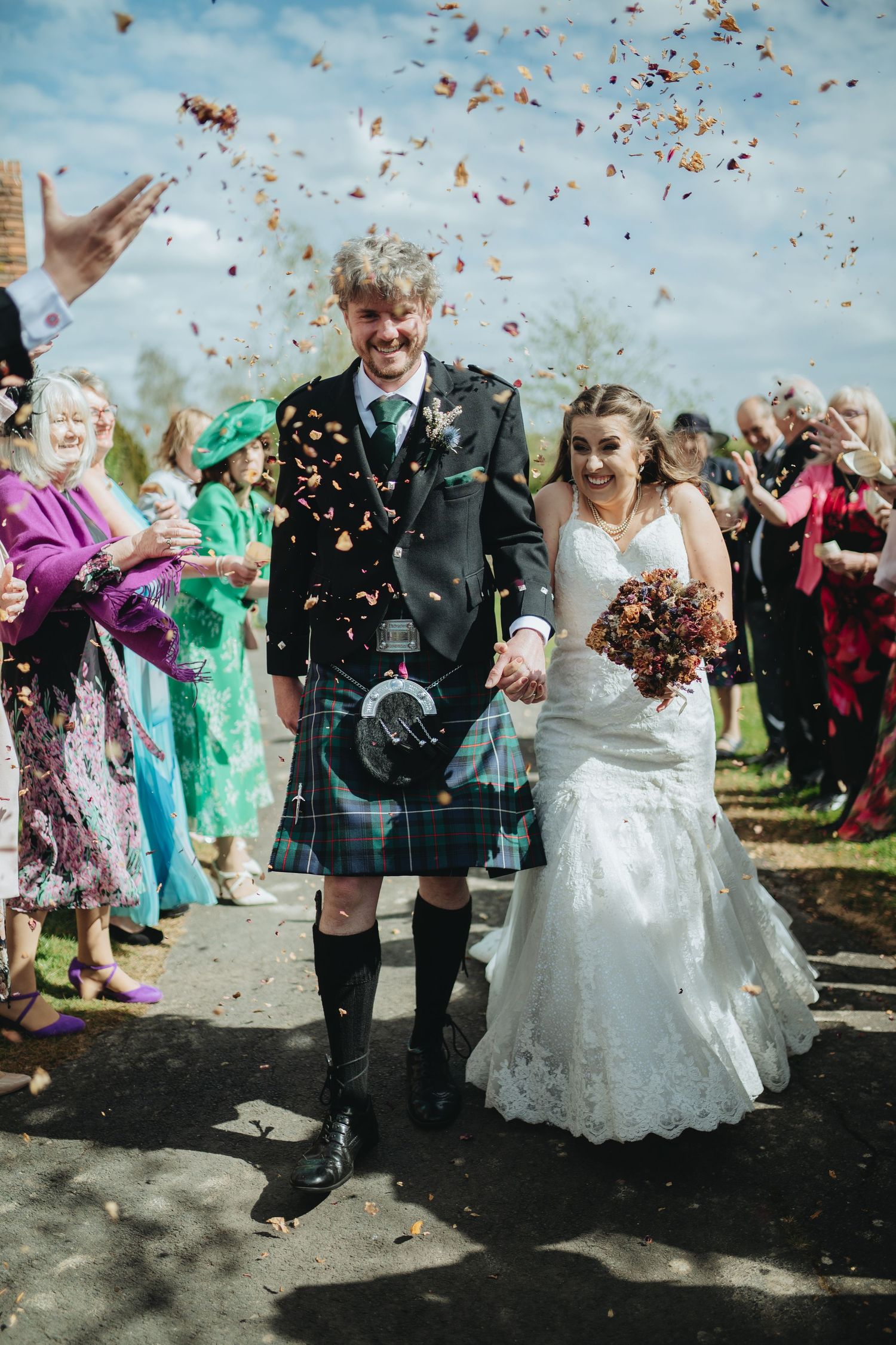 Bride and groom walk through confetti shower, groom in tartan kilt, bride in lace gown, guests celebrating outdoors.