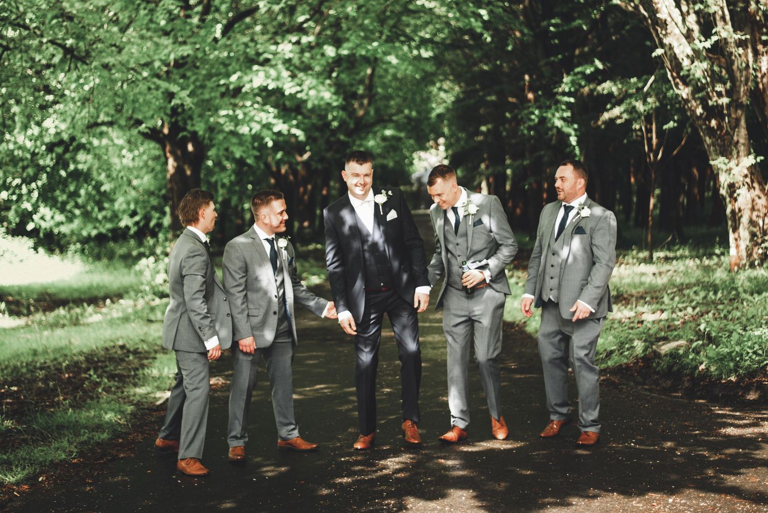 Groomsmen in matching gray suits and brown shoes laughing together in a forest setting.