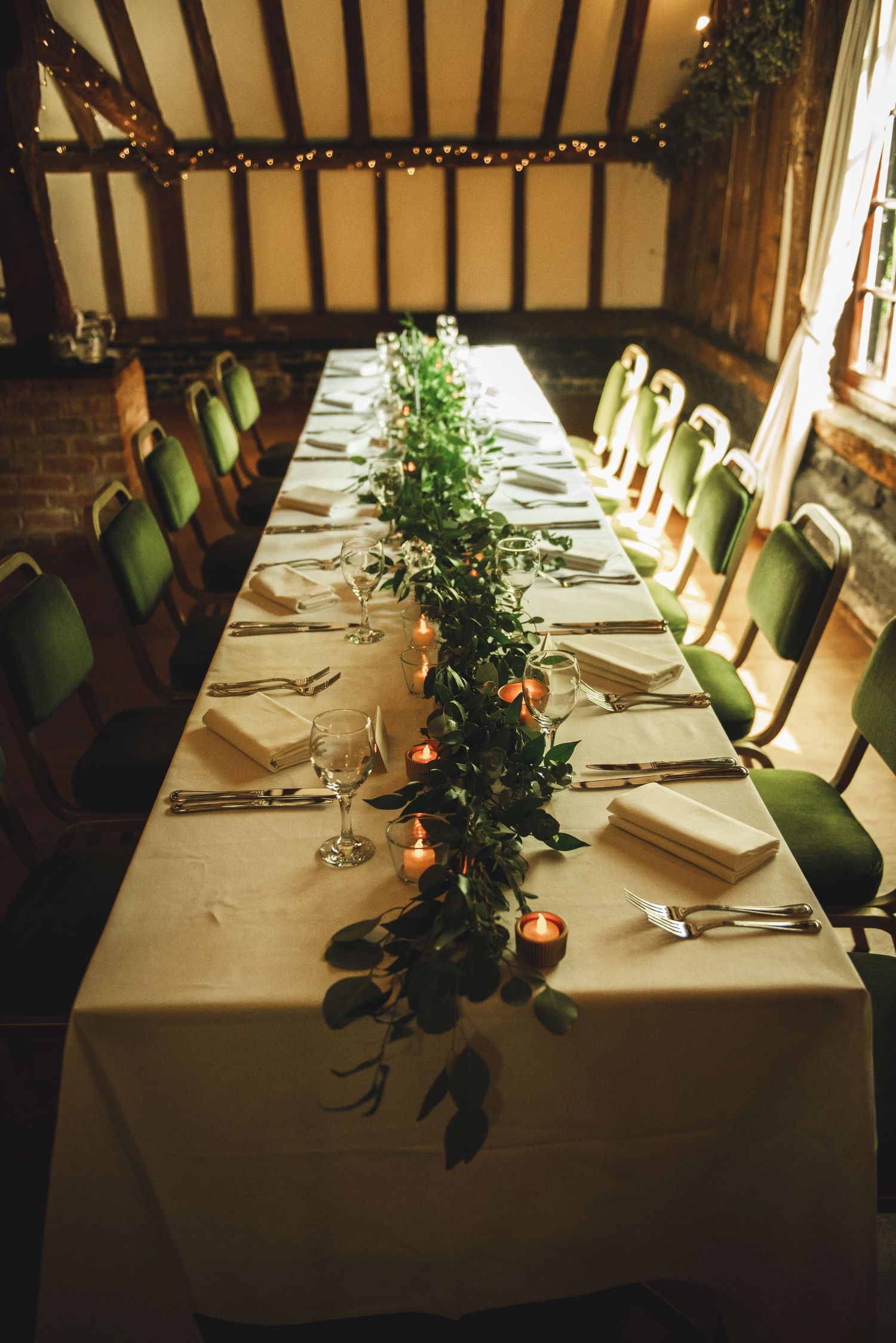 The Plough in Leigh KenLong dining table with white linens, green chairs, and a natural garland centerpiece in an elegant wood-beamed venue.
