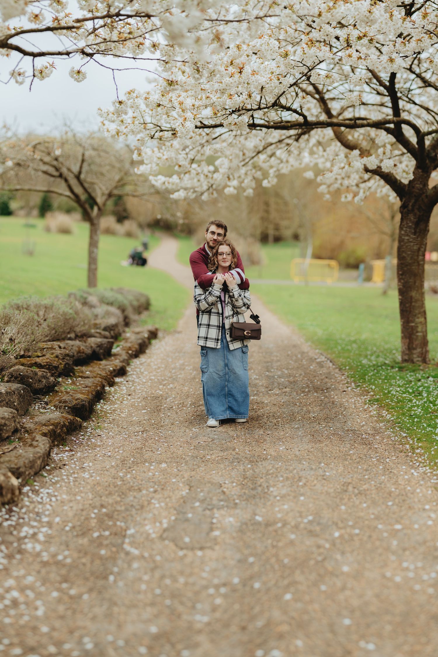 Couple embracing on a tree-lined gravel path with white cherry blossoms in bloom during spring.