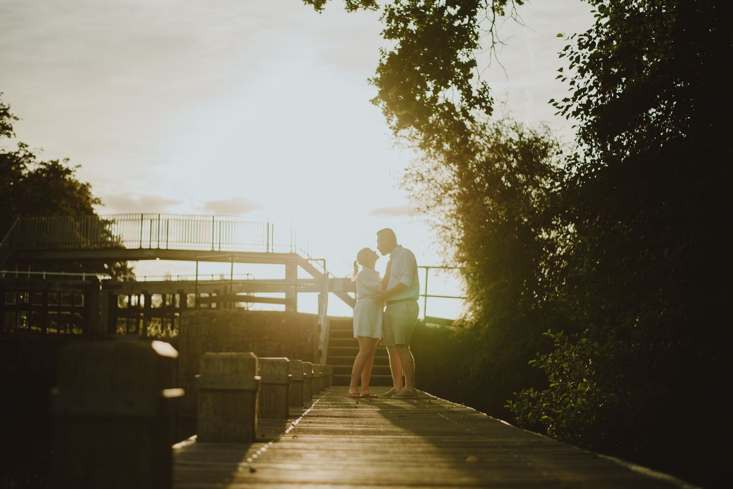 Silhouetted couple embraces on wooden dock during golden sunset with trees framing the romantic scene.