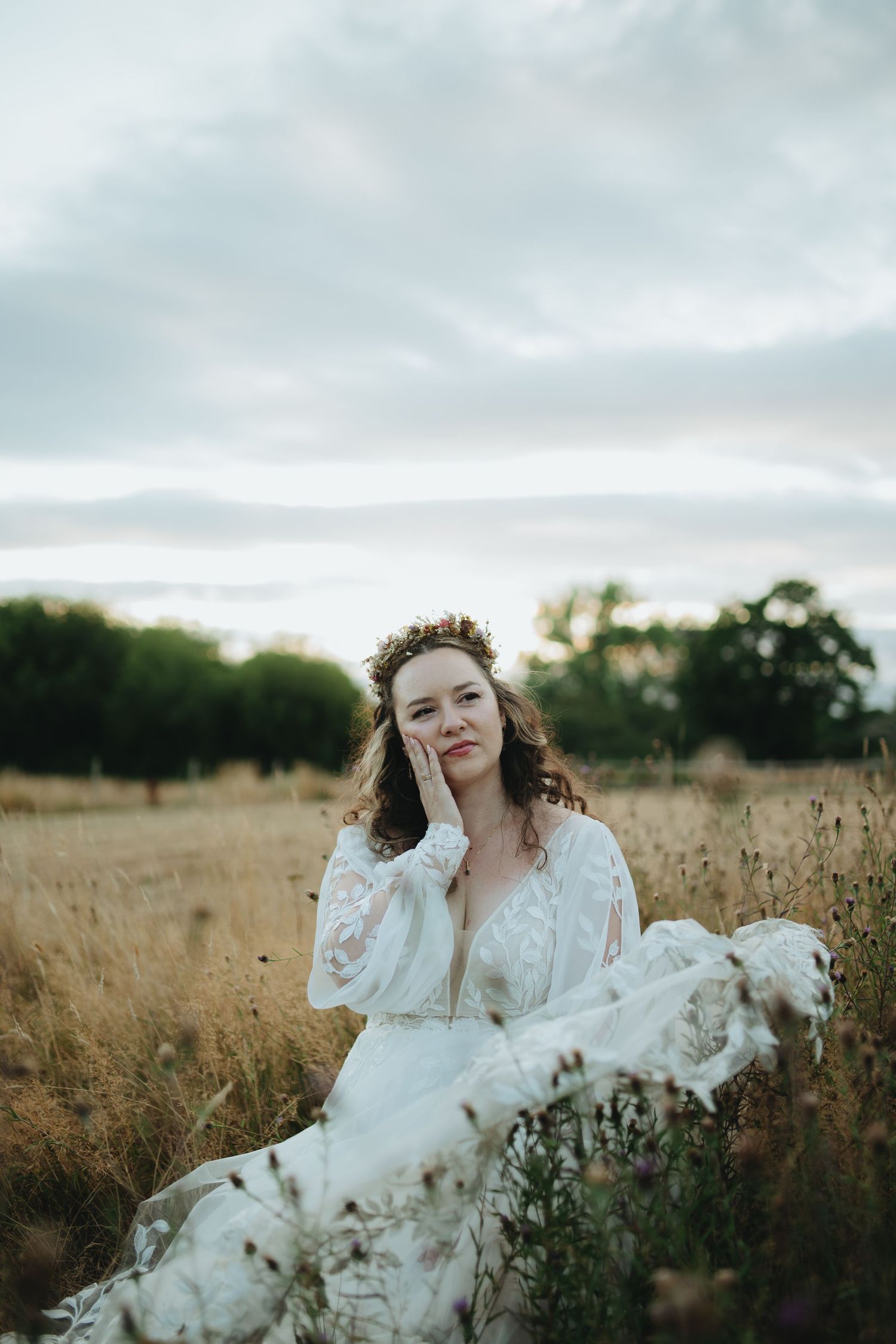 A bride in a white lace gown and floral crown sits gracefully in a golden field under a cloudy sky.