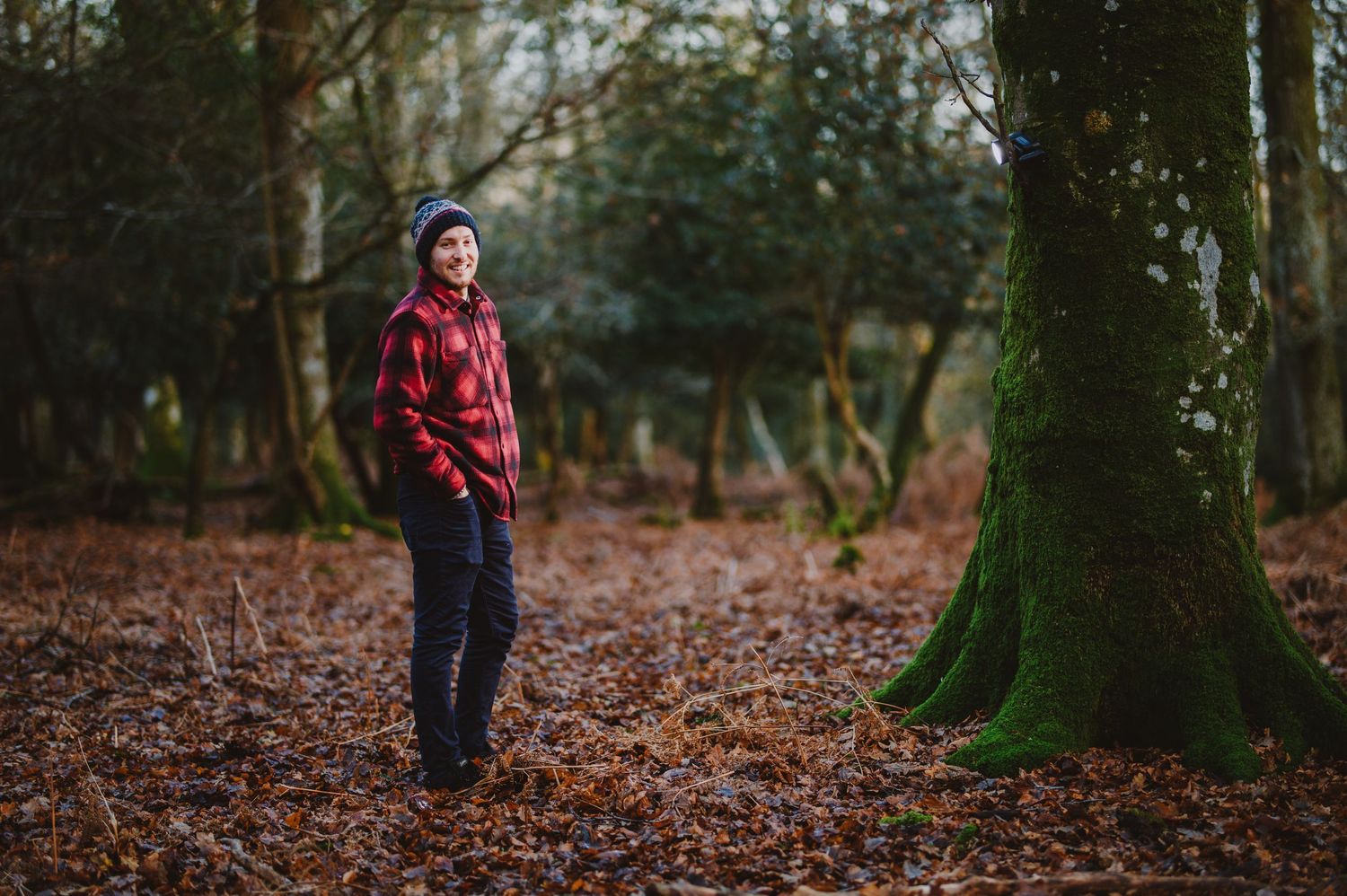 Person in red plaid shirt and dark pants walks through autumn forest with fallen leaves on ground.