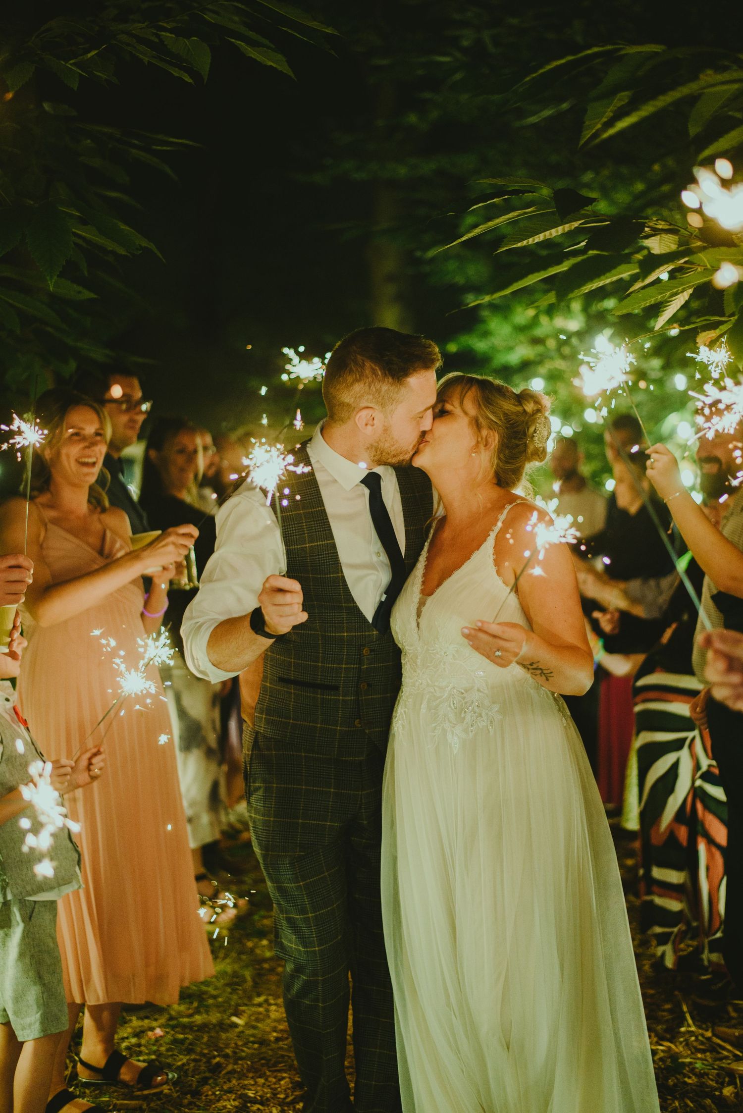 Wedding sparklers illuminate the evening as couples dance under lush green trees at an outdoor reception.