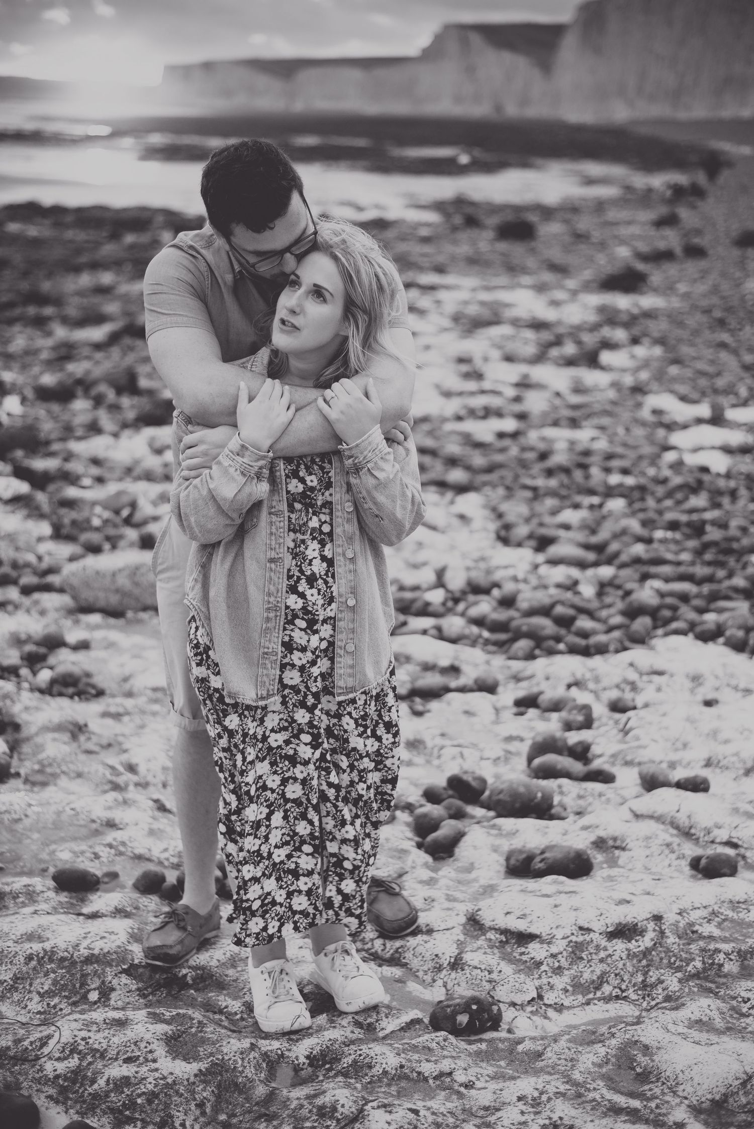 Black and white portrait of a couple sharing a tender moment on a rocky beach near coastal cliffs.