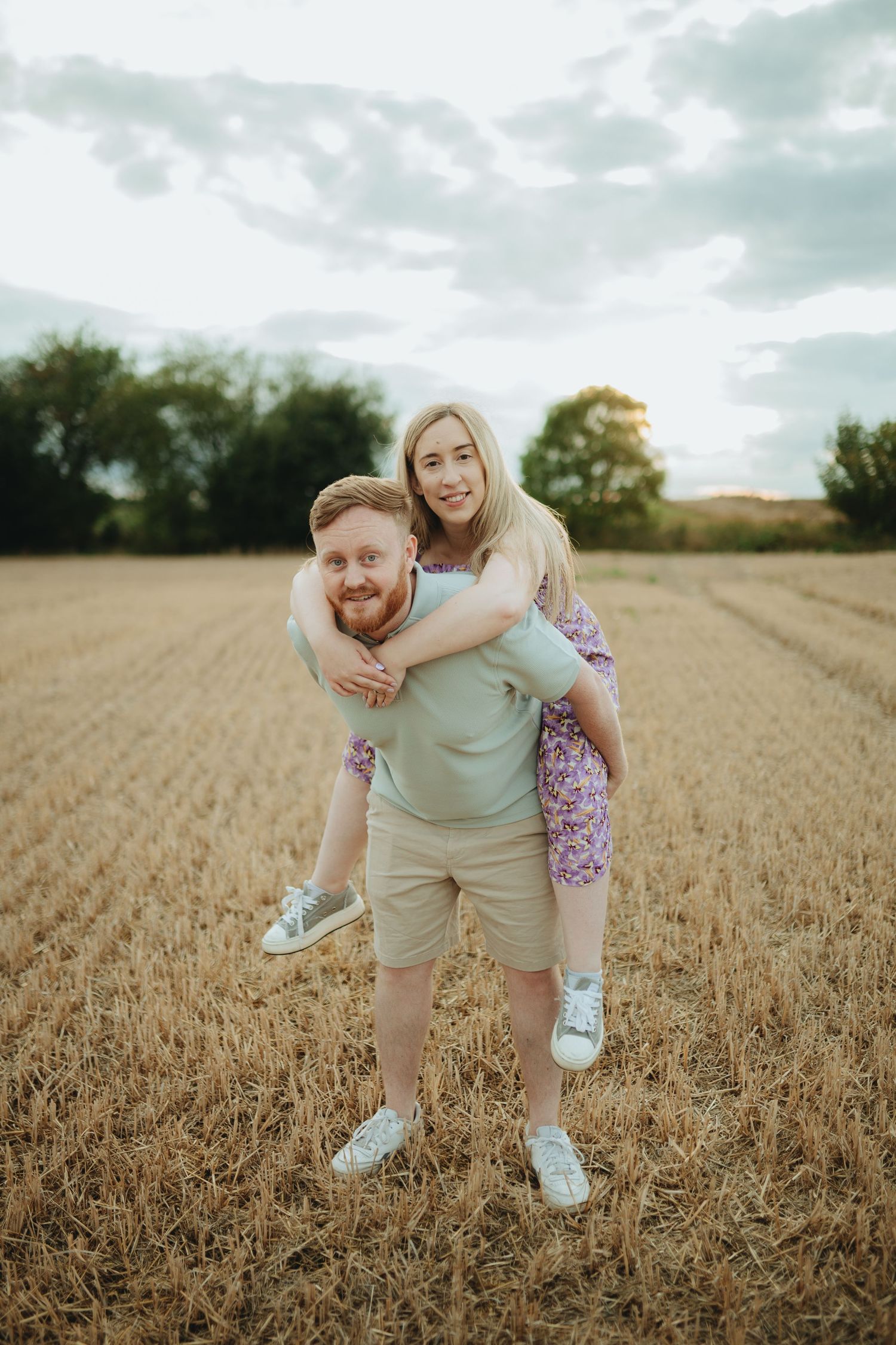 A man with a red beard gives a piggyback ride to a blonde woman in a harvested wheat field under a cloudy sky.