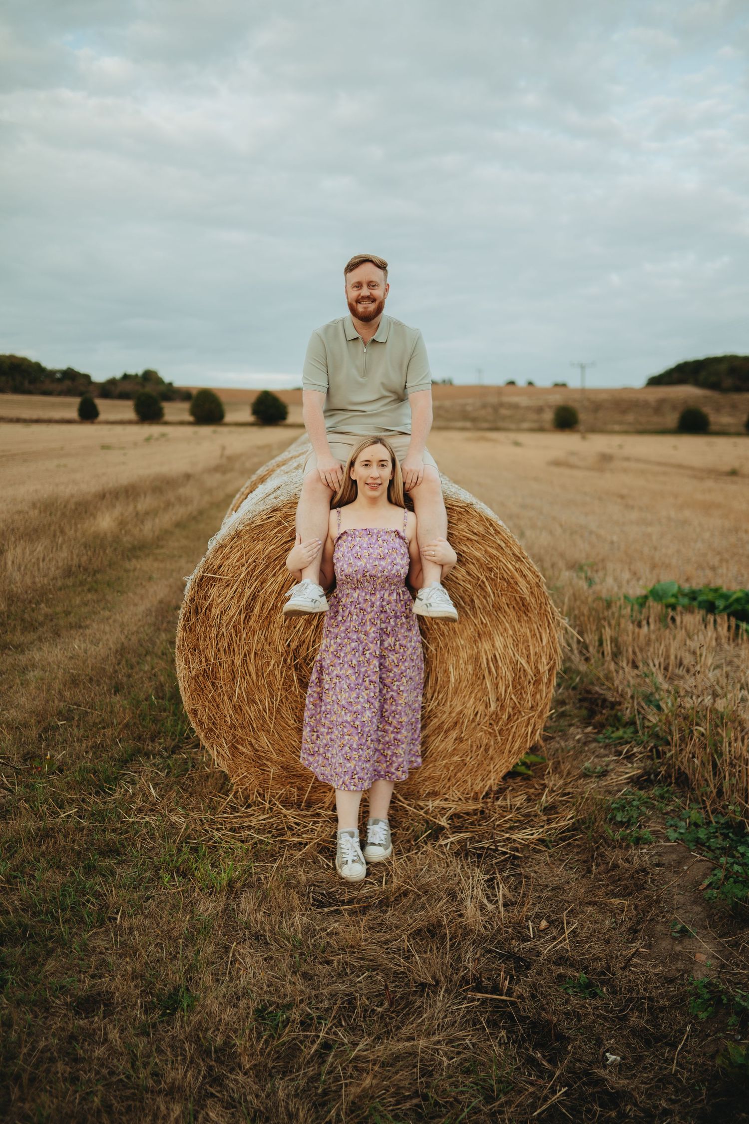 Couple posing with hay bale in harvested field, man sitting on top, woman standing in floral dress.