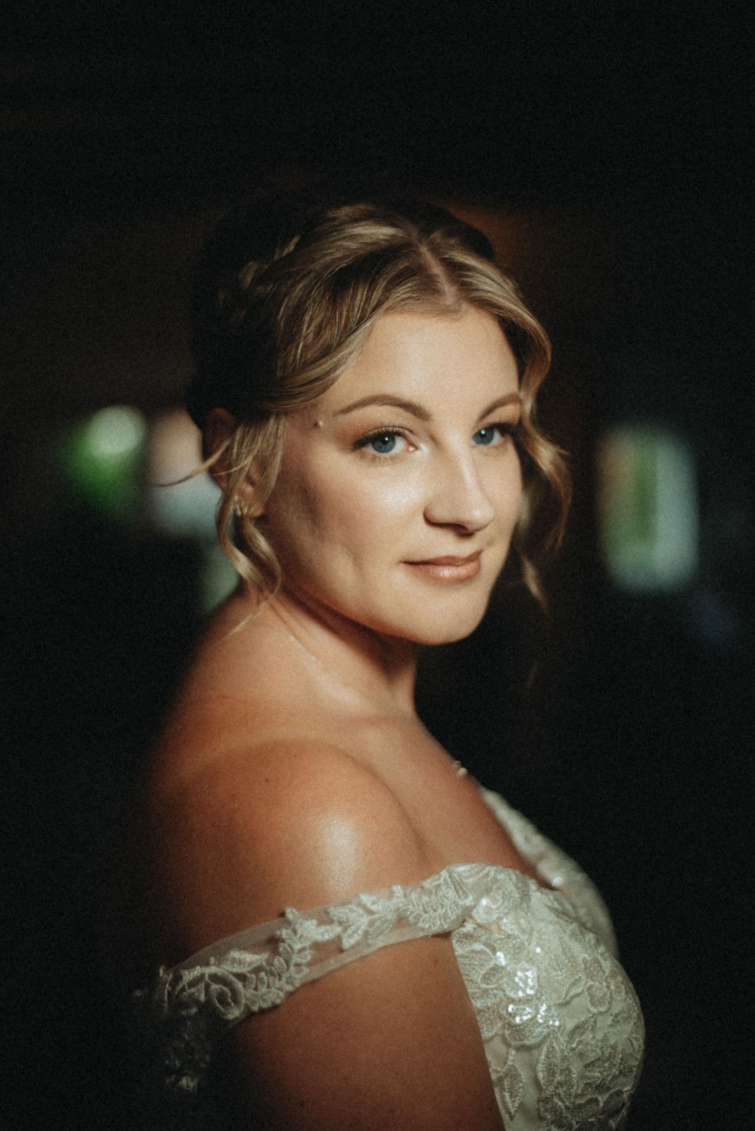A bride in an off-shoulder lace gown poses elegantly with soft lighting highlighting her updo and delicate jewelry.