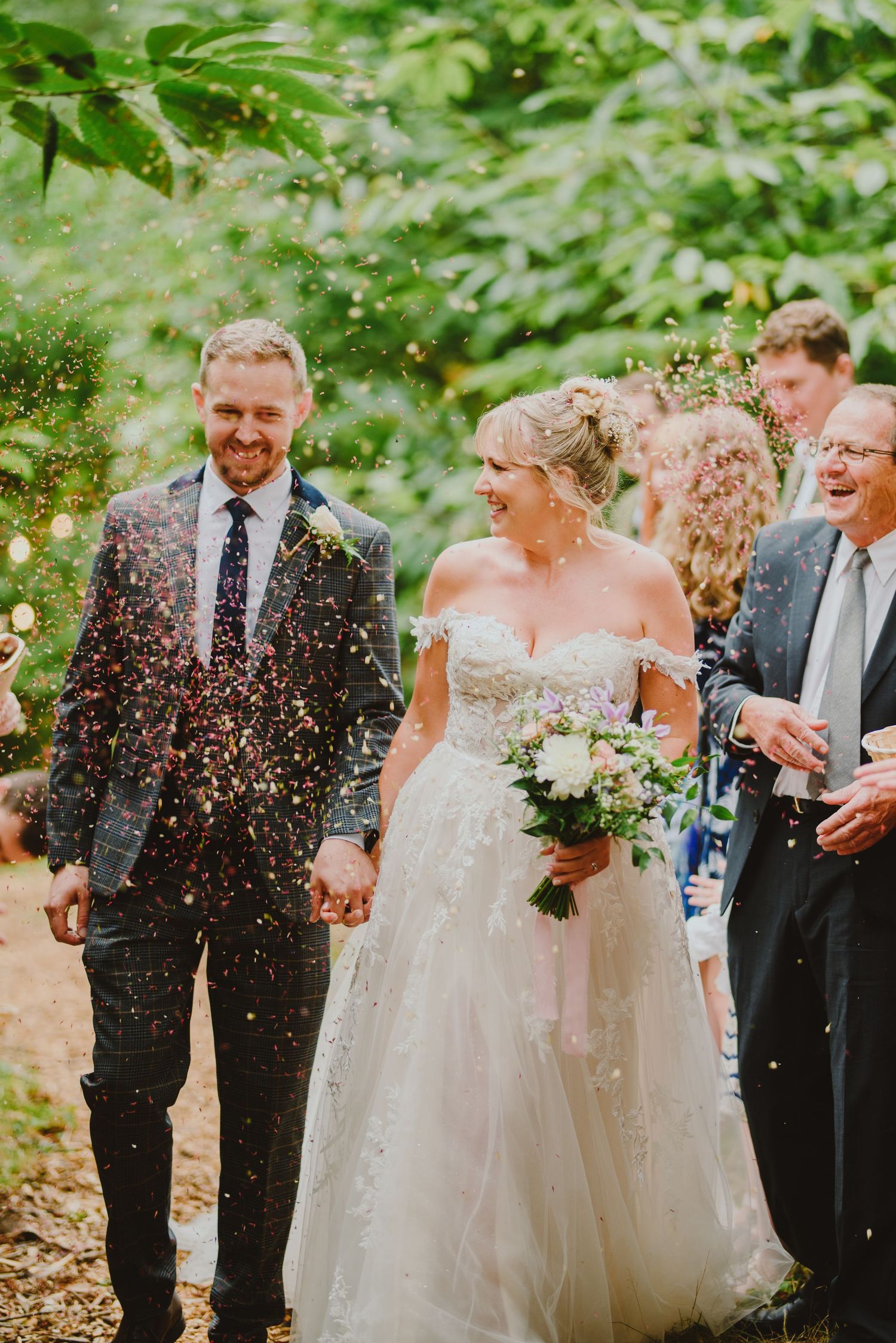 Wedding party sprinkled with confetti during outdoor celebration in lush green garden setting.