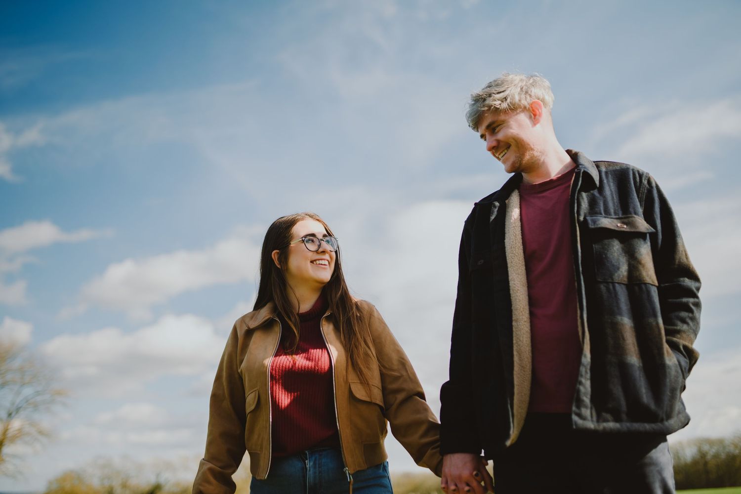 A couple stands together in a field during golden hour with blue skies and clouds in the background.