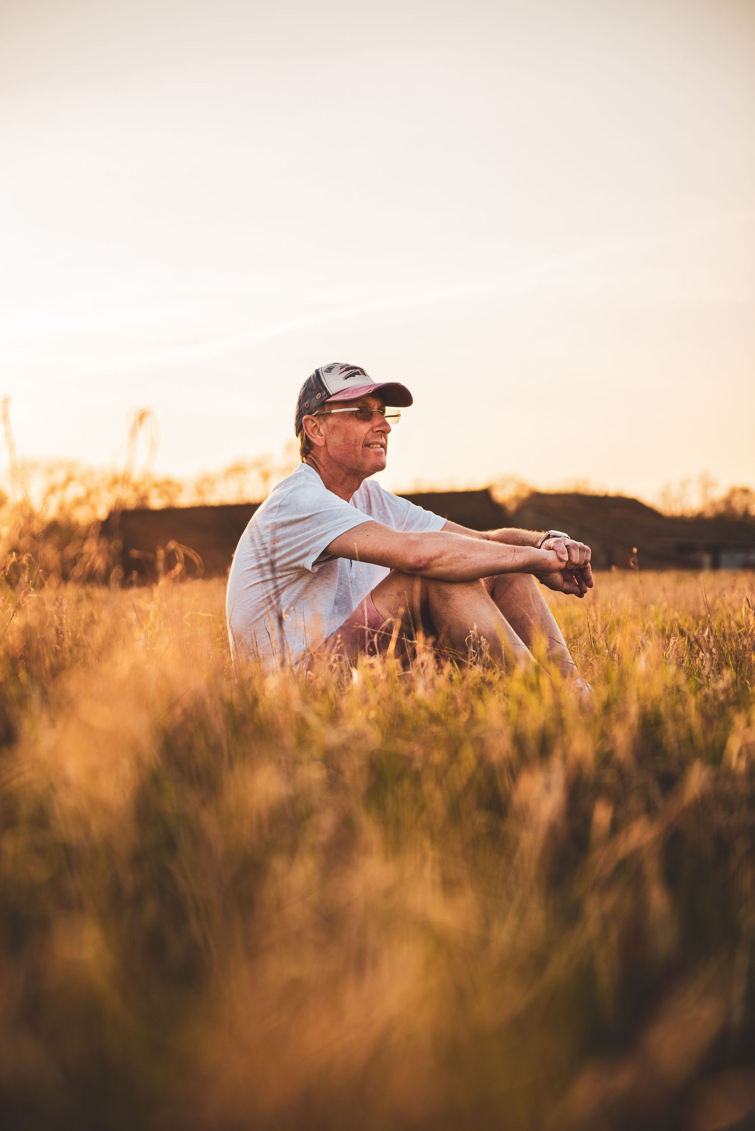 A person sits peacefully in a golden wheat field at sunset wearing a baseball cap and white shirt.