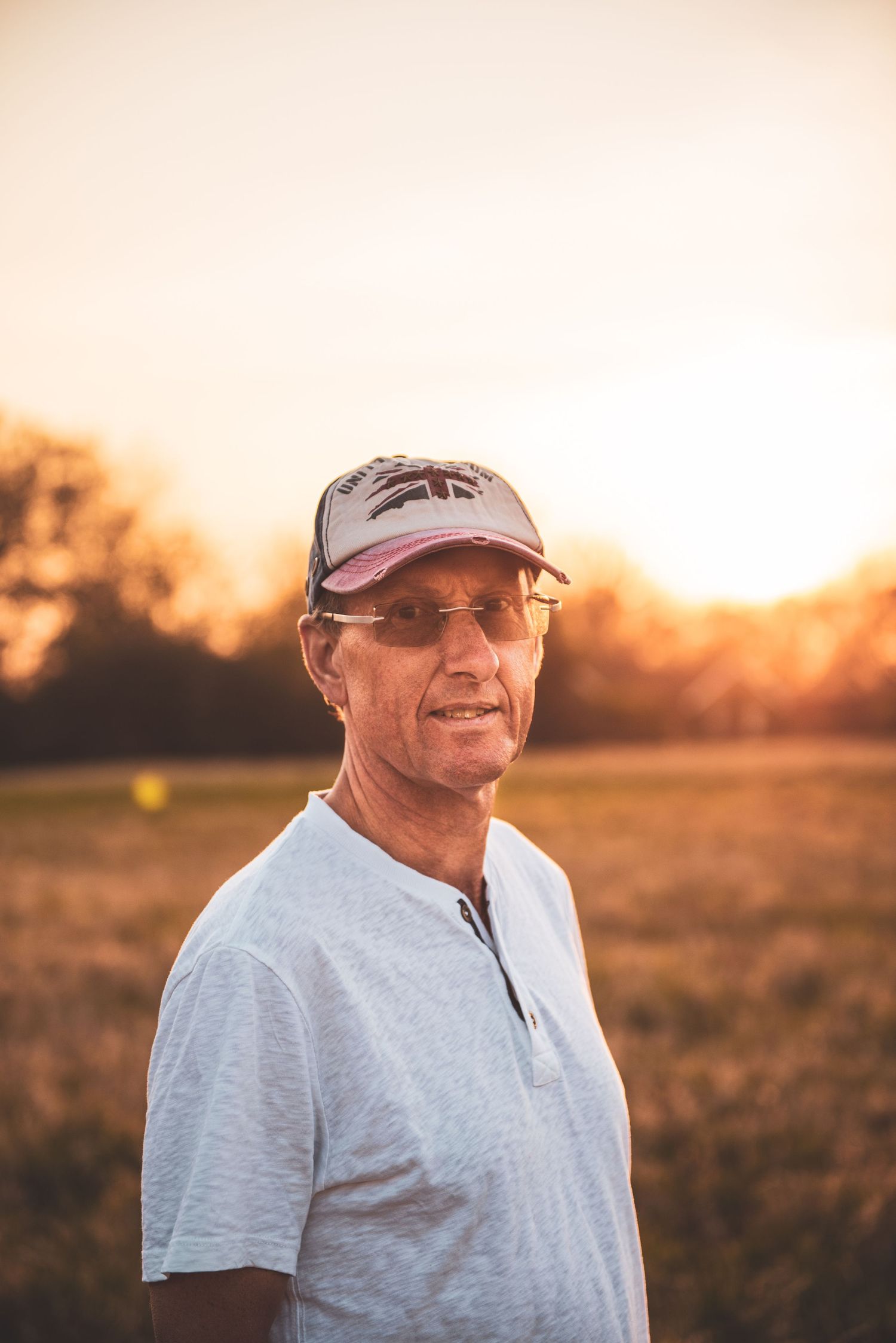 Person in grey t-shirt and baseball cap smiling outdoors against a golden sunset sky and grassy field.