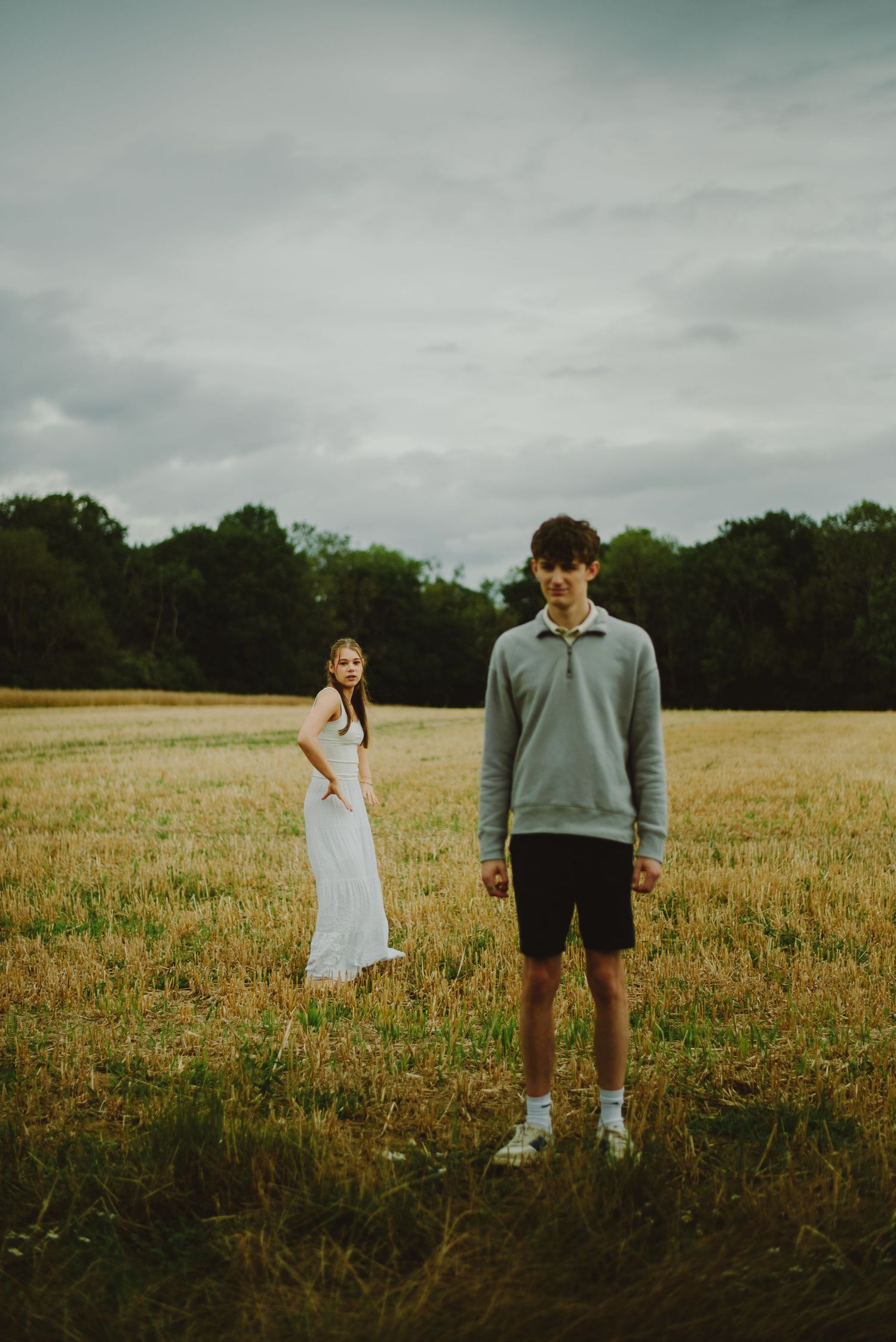 Two people stand apart in a golden field on an overcast day with dark trees in the background.