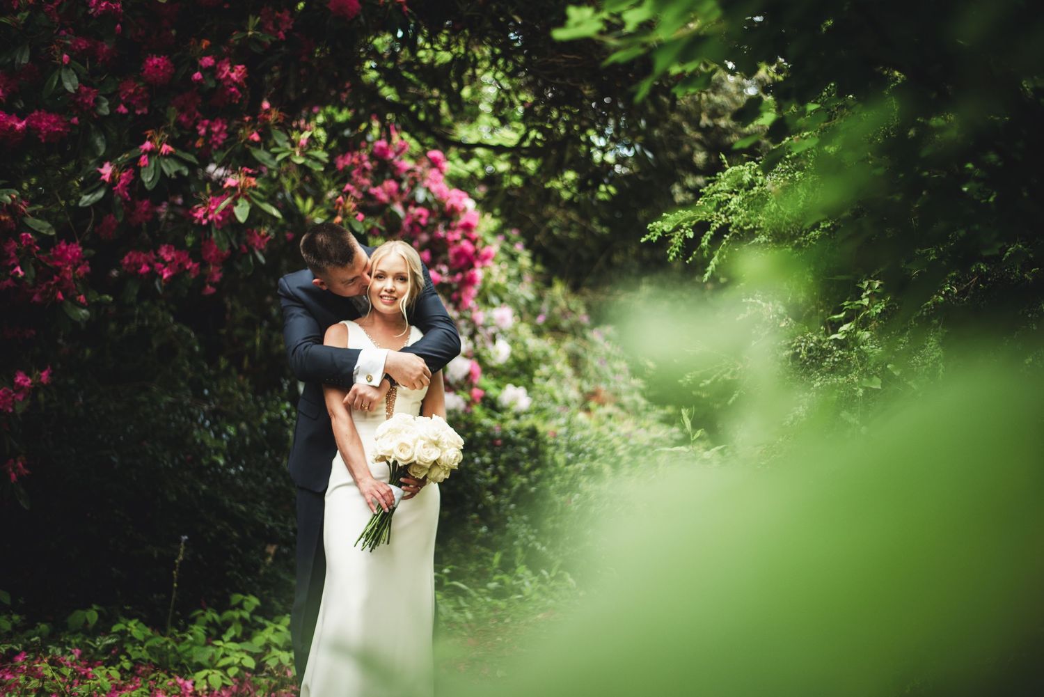 East Sussex wedding. A romantic wedding moment captured in a garden setting with vibrant pink flowers and lush greenery in the background.