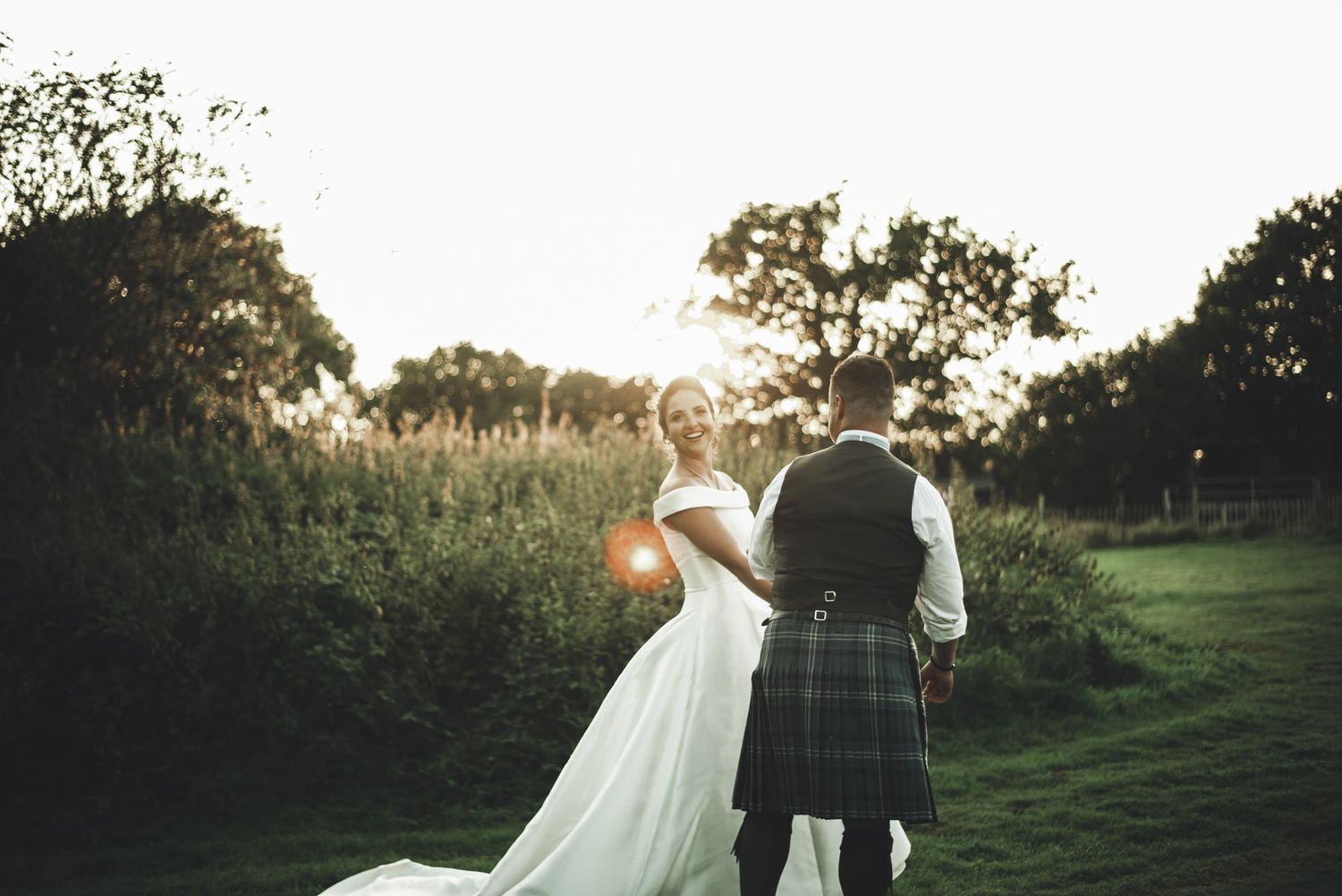 A couple walks together through a sunlit field during golden hour, with the bride's veil flowing in the breeze.