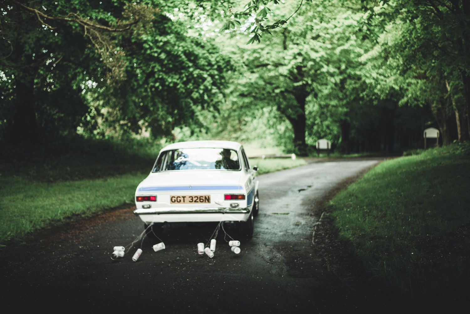 Vintage white car parked on a wet forest road surrounded by lush green trees.