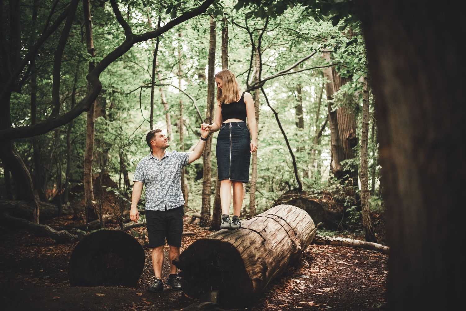 A couple balances on a fallen log in a lush forest during an engagement photoshoot.