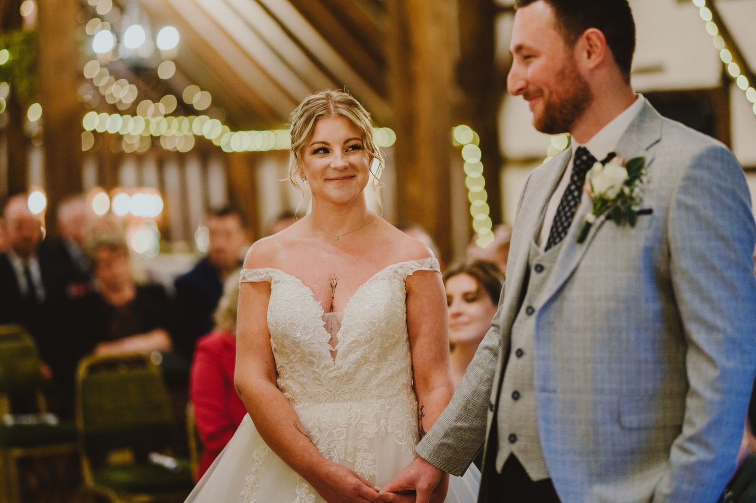 A couple stands together during their wedding ceremony in a rustic barn venue with glowing lights overhead.