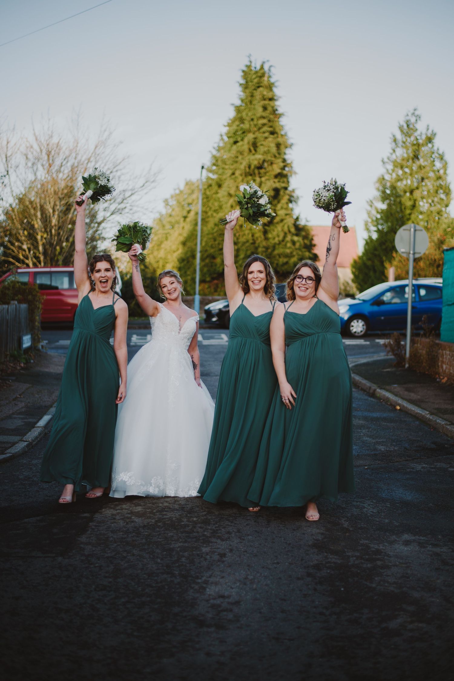 A group of bridesmaids in emerald green dresses pose playfully with the bride in her white gown outdoors.