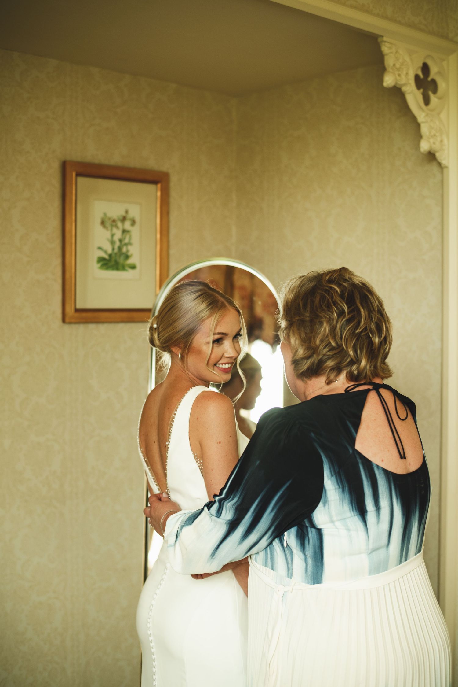 Bride getting dressed in elegant white gown while looking in mirror with helper.