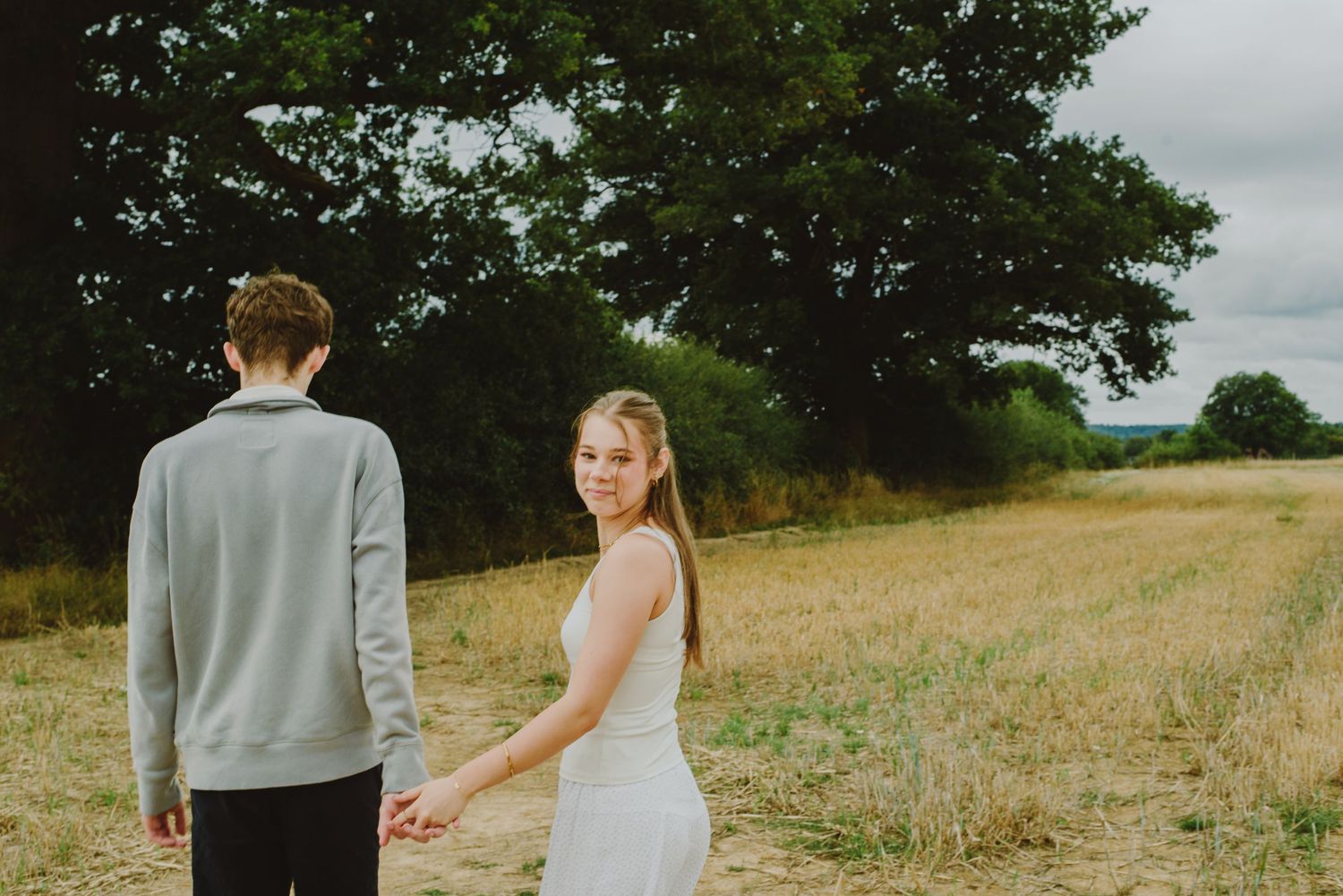 A couple holds hands while walking through a golden grass field on an overcast day.