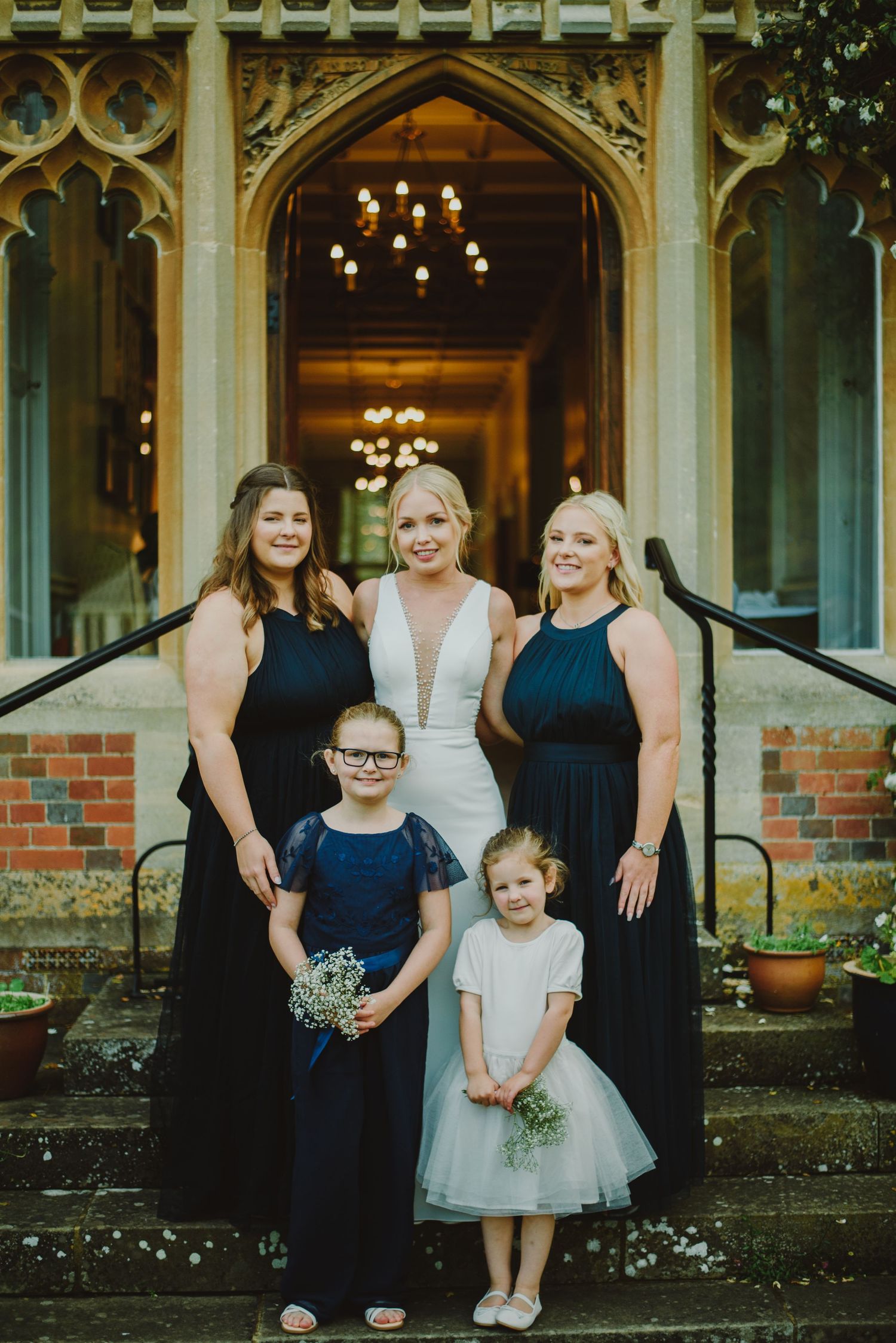 Wedding party in navy blue dresses poses on stone steps of elegant historic building with chandeliers visible inside.