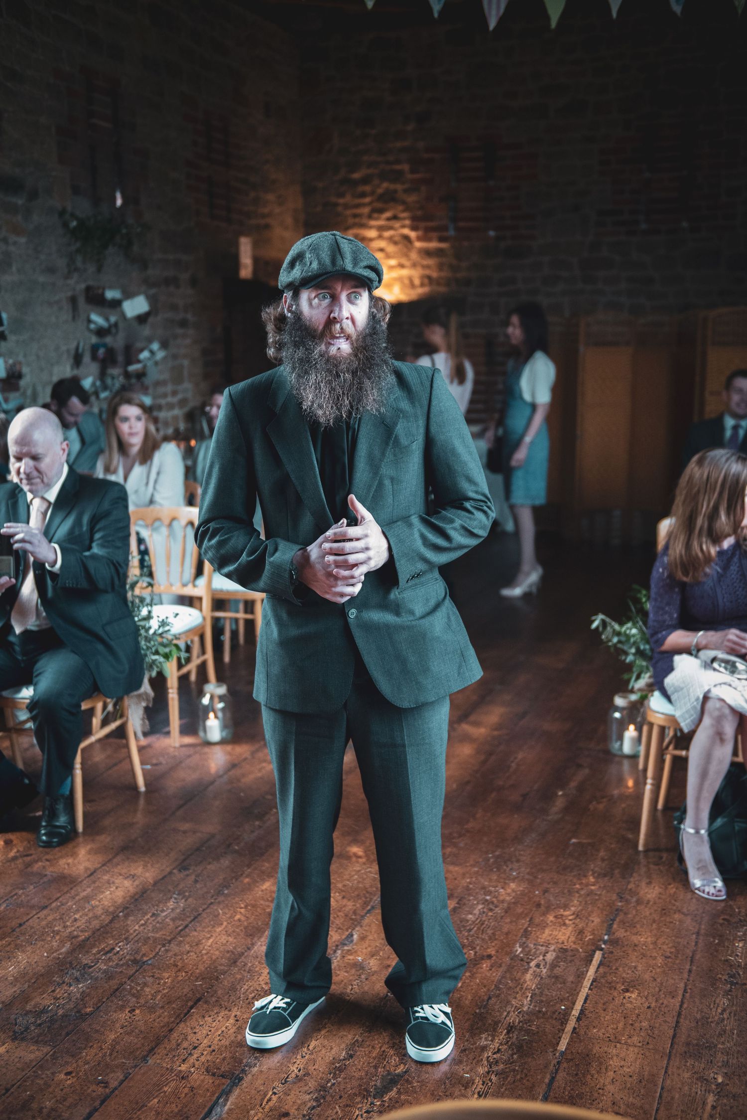 Guest in green suit and flat cap stands on wooden floor at indoor wedding reception.