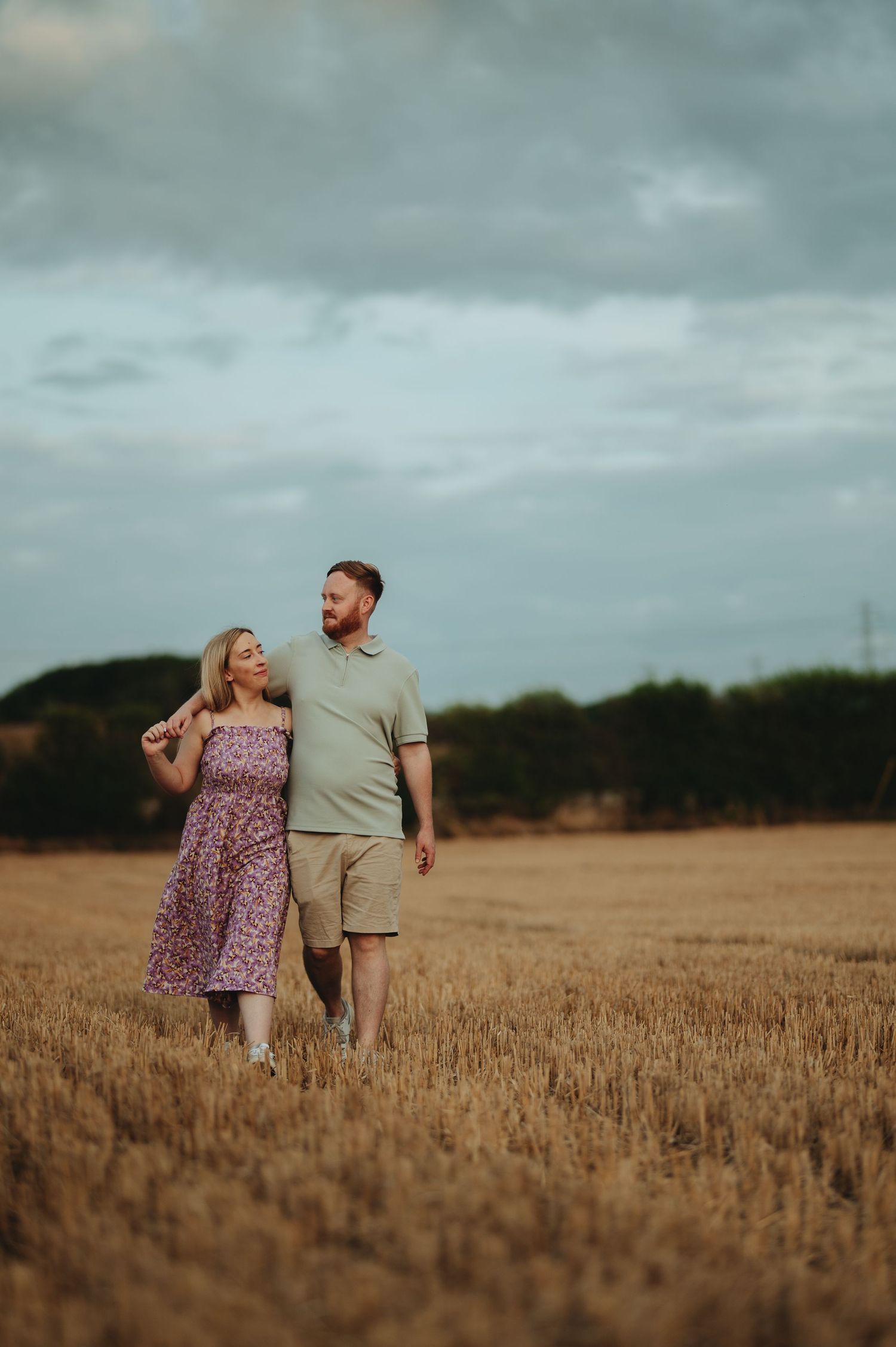 A couple walks hand-in-hand through a golden wheat field under a cloudy sky, the woman in a floral dress and man in casual shorts.