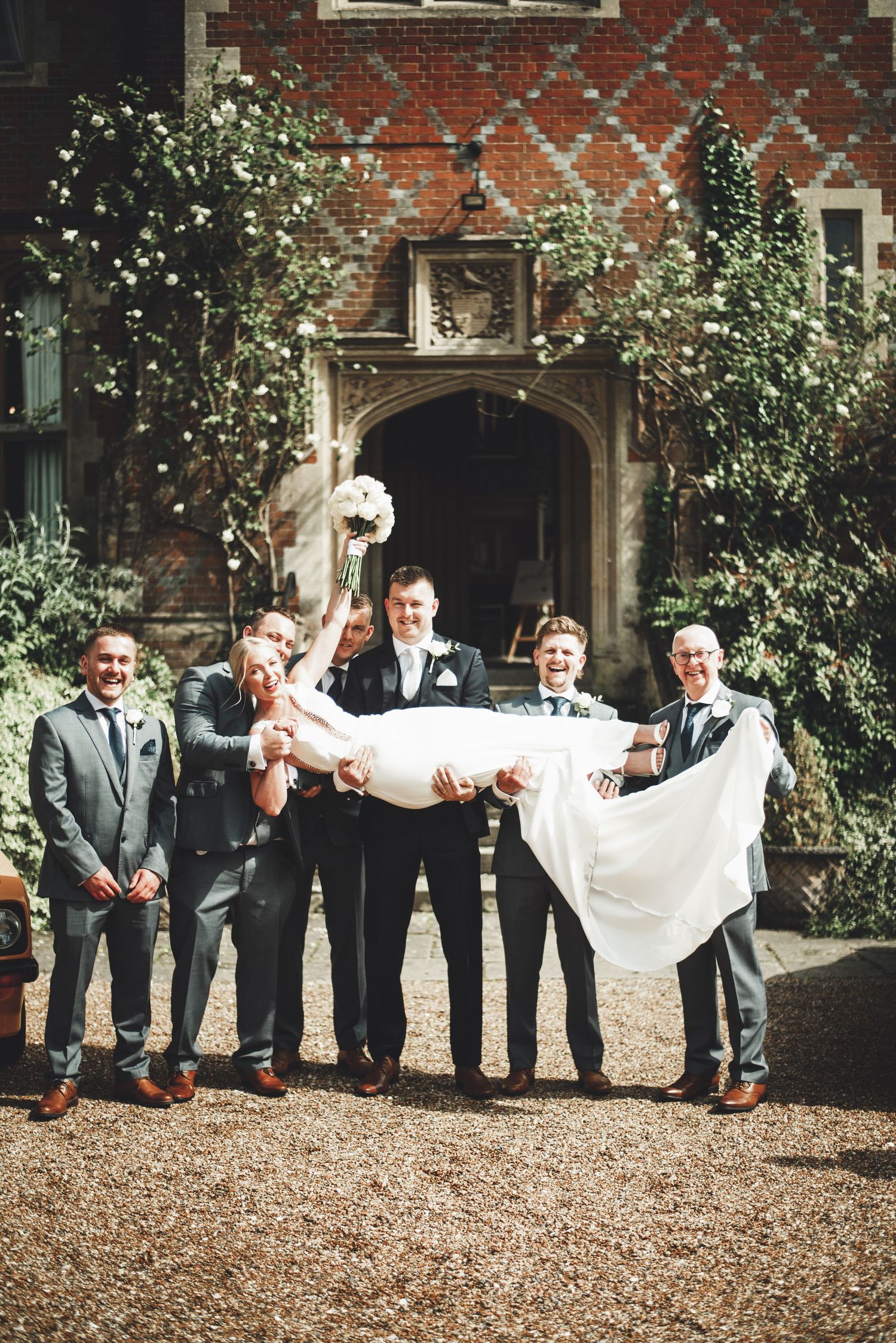 Wedding party in formal attire lifting couple in front of historic brick building entrance.