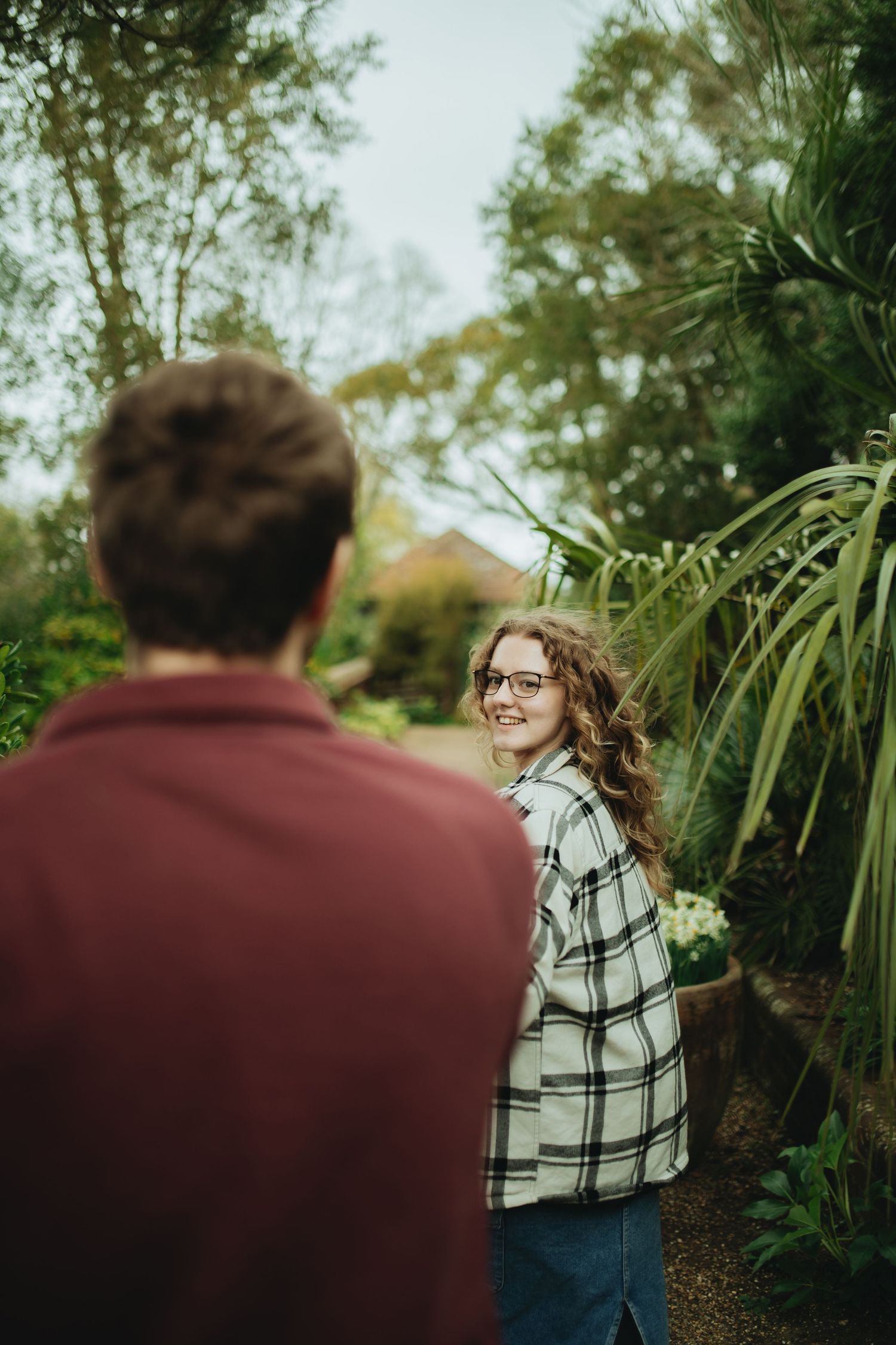 A smiling young woman in glasses and plaid shirt glances back at a man on a lush garden path.