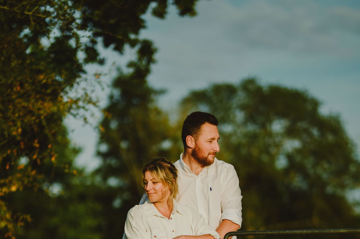 Couple wearing light colored outfits smile and embrace during open air photography session in wooded setting.