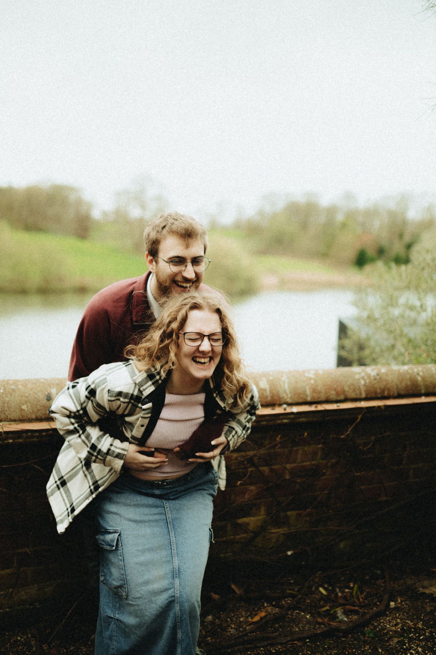 A laughing couple embraces on a stone bridge overlooking a serene lake, woman in plaid shirt and denim skirt.